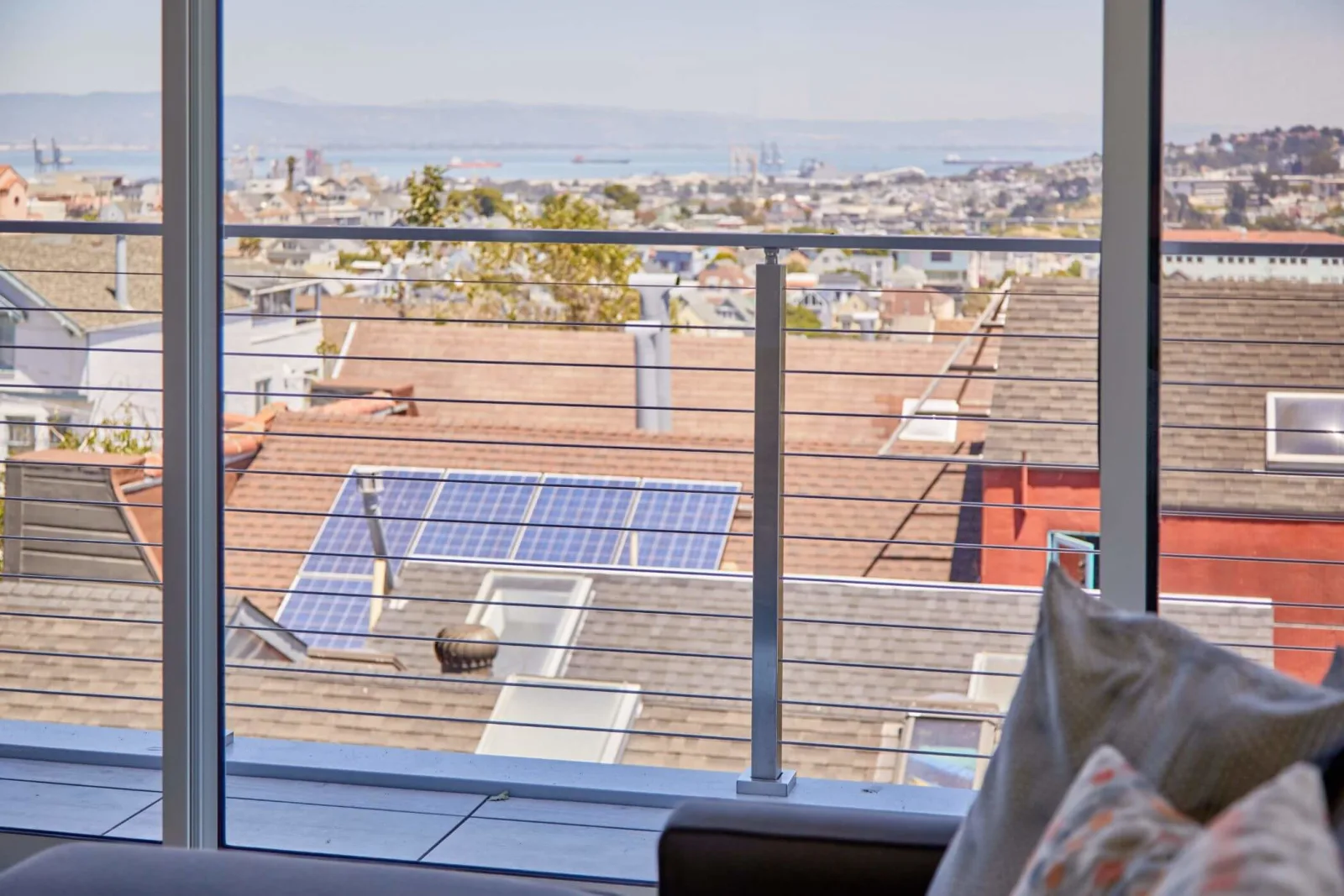 City rooftops and solar panels seen past a balcony railing from an interior seat