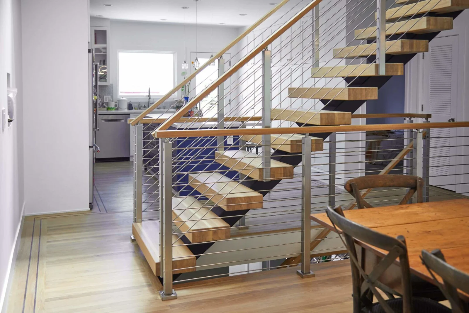 Stair treads and rod railing seen across a dining area toward a bright kitchen