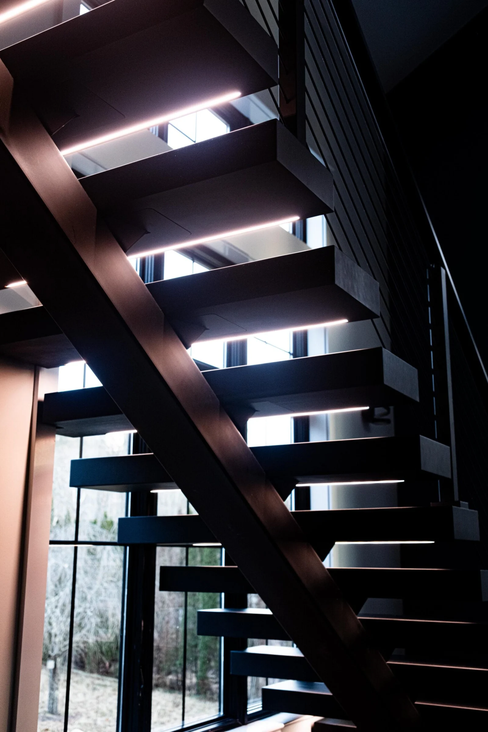 Underlit treads glow along a black stringer stair against tall dark windows at night