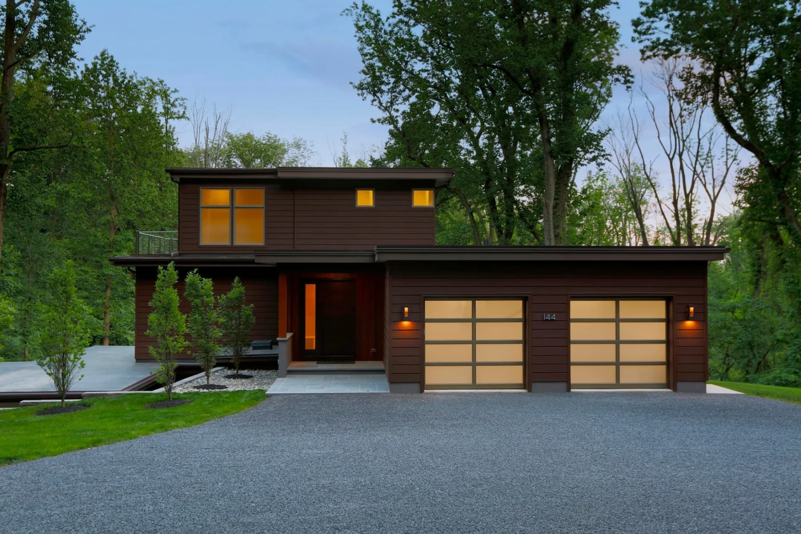 Dark-clad two-level house at dusk with lit garage doors and gravel drive