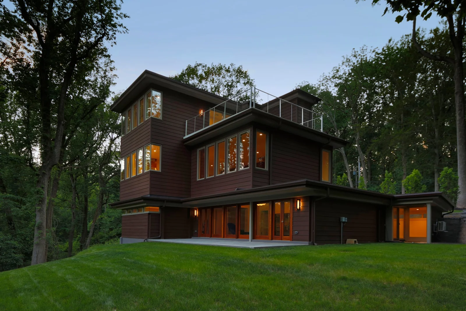 Rear of the dark-clad house at dusk with lit windows above a lawn