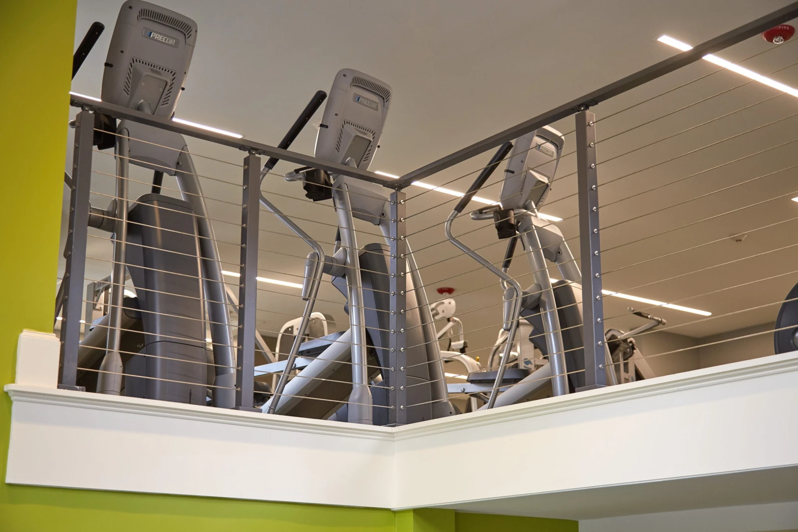 Mezzanine cable railing guarding elliptical machines above a green painted wall
