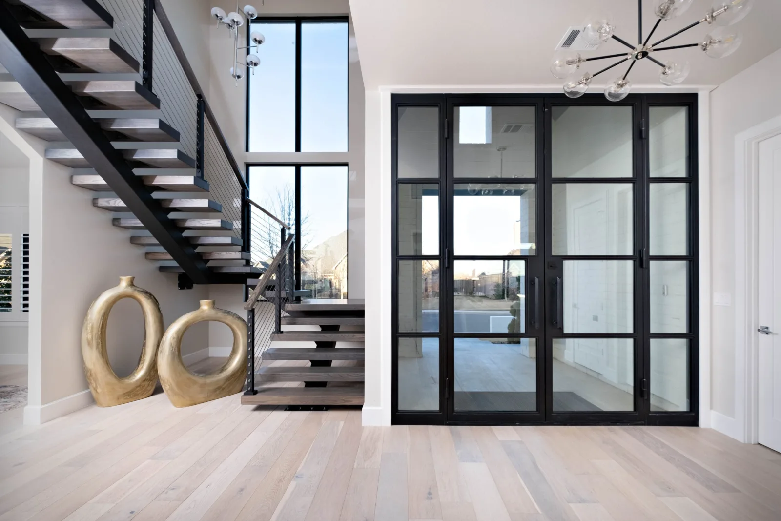 Black stair with light treads and rod railing rising beside a glass-paneled entry with gold vessels