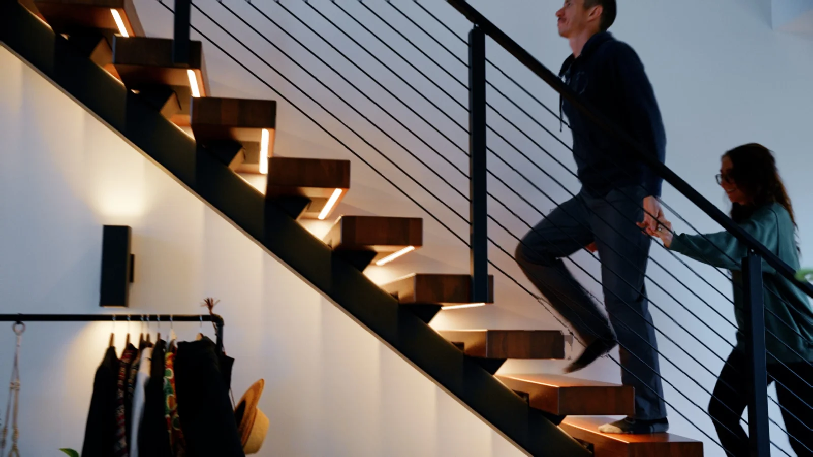 Two people climbing an open-riser flight with lit treads behind black rod railing