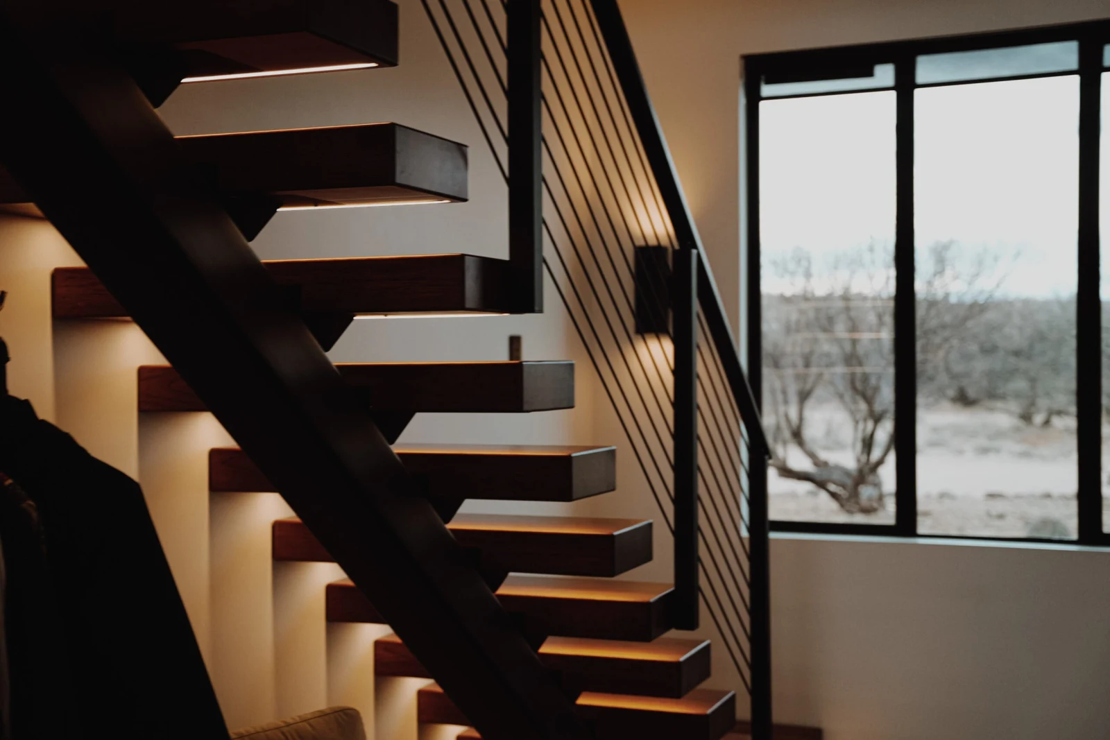Floating stair beside a tall window that looks out onto dry brush and open ground