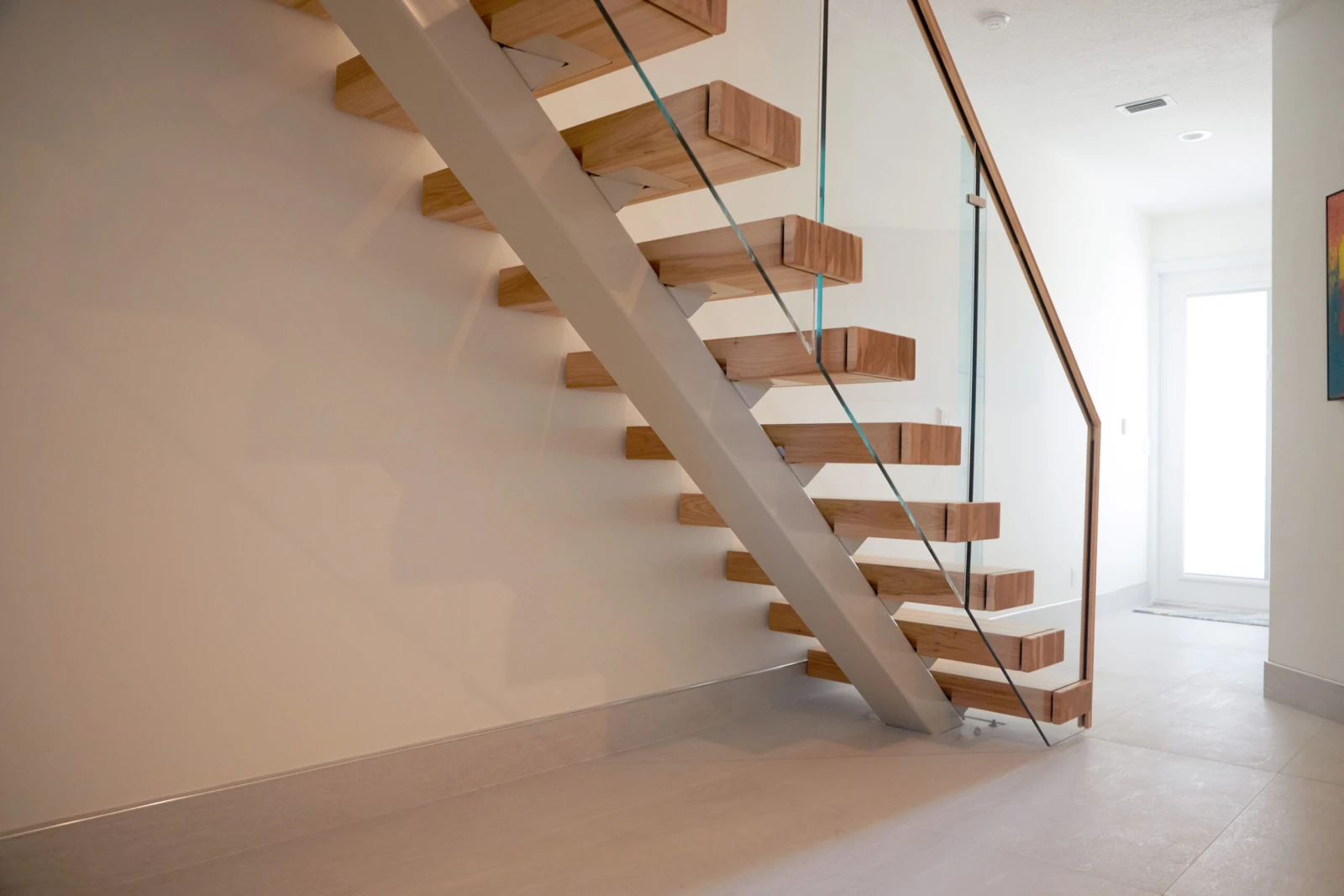 Wood treads on a pale center stringer with frameless glass panels along a quiet tiled hallway