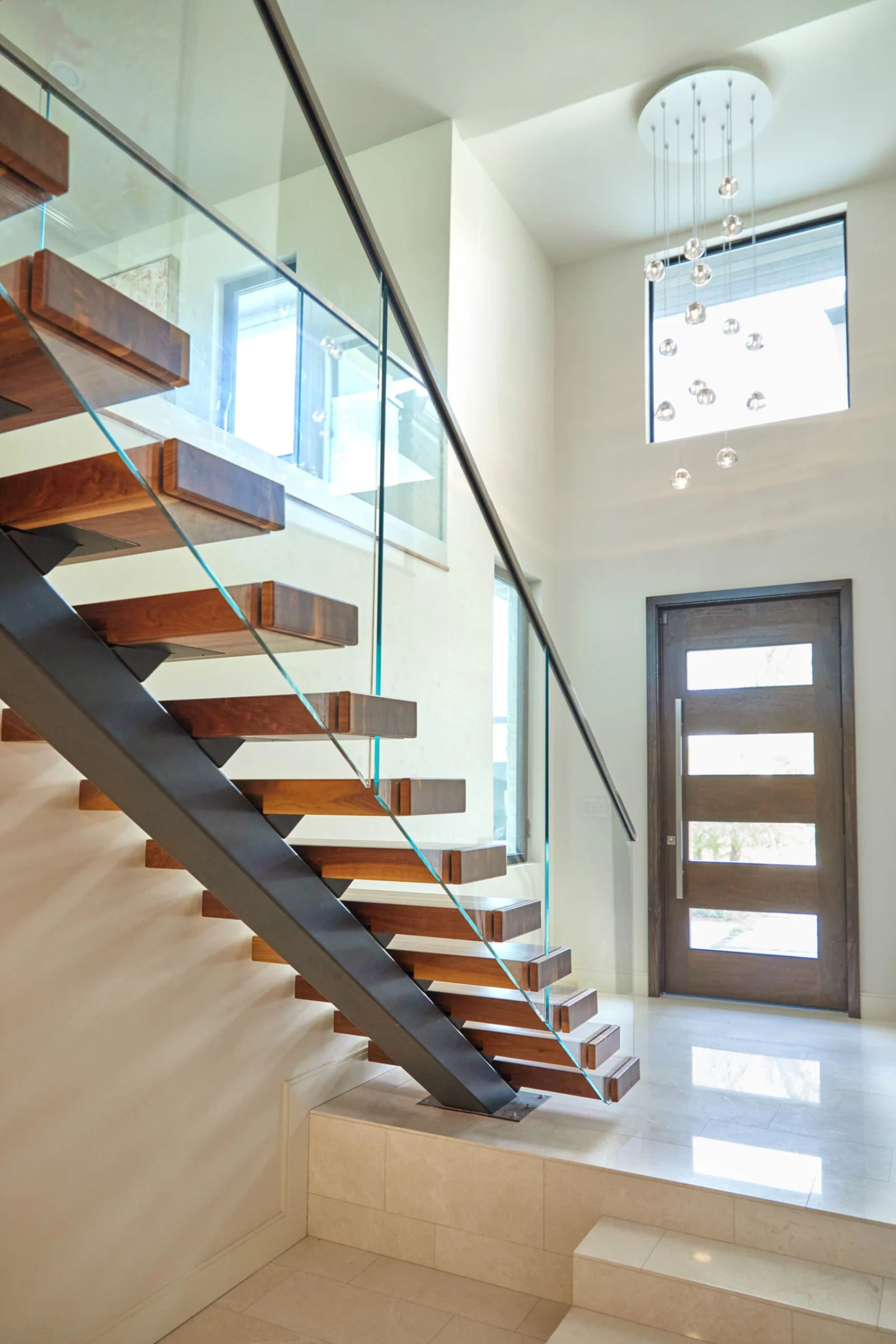 Entry foyer stair with glass railing beside the front door and chandelier