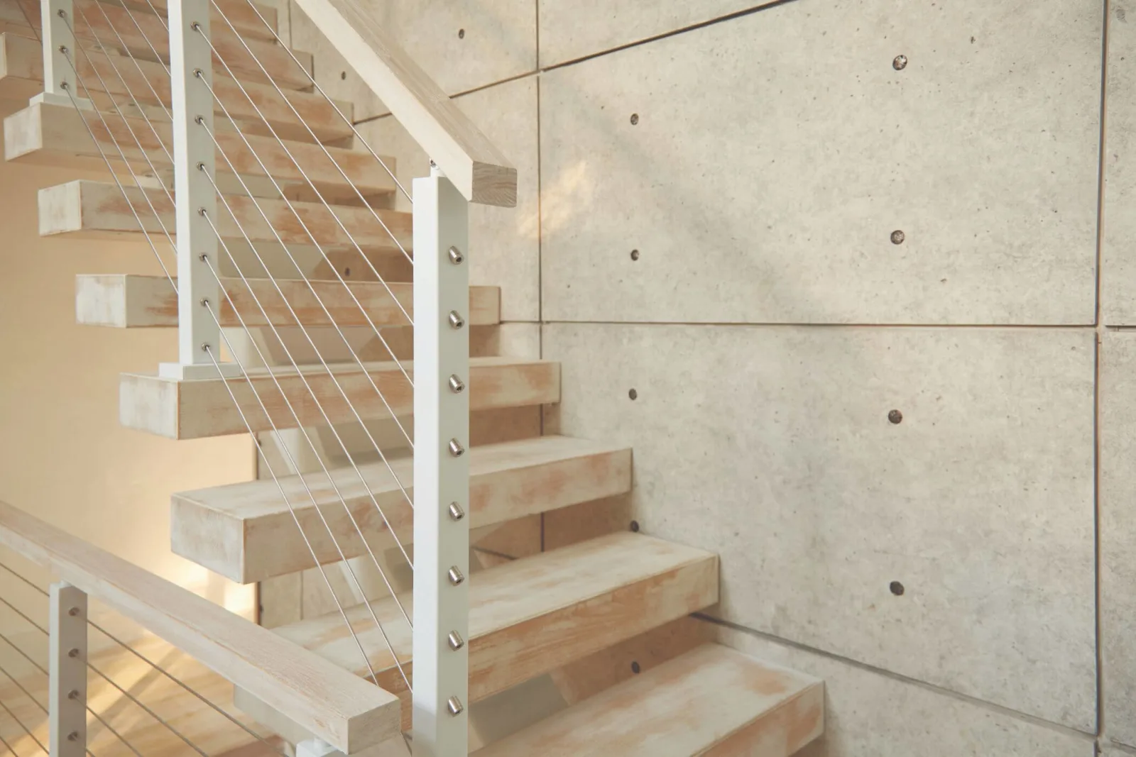 Floating stair with pale wood treads and cable railing rising past a concrete wall