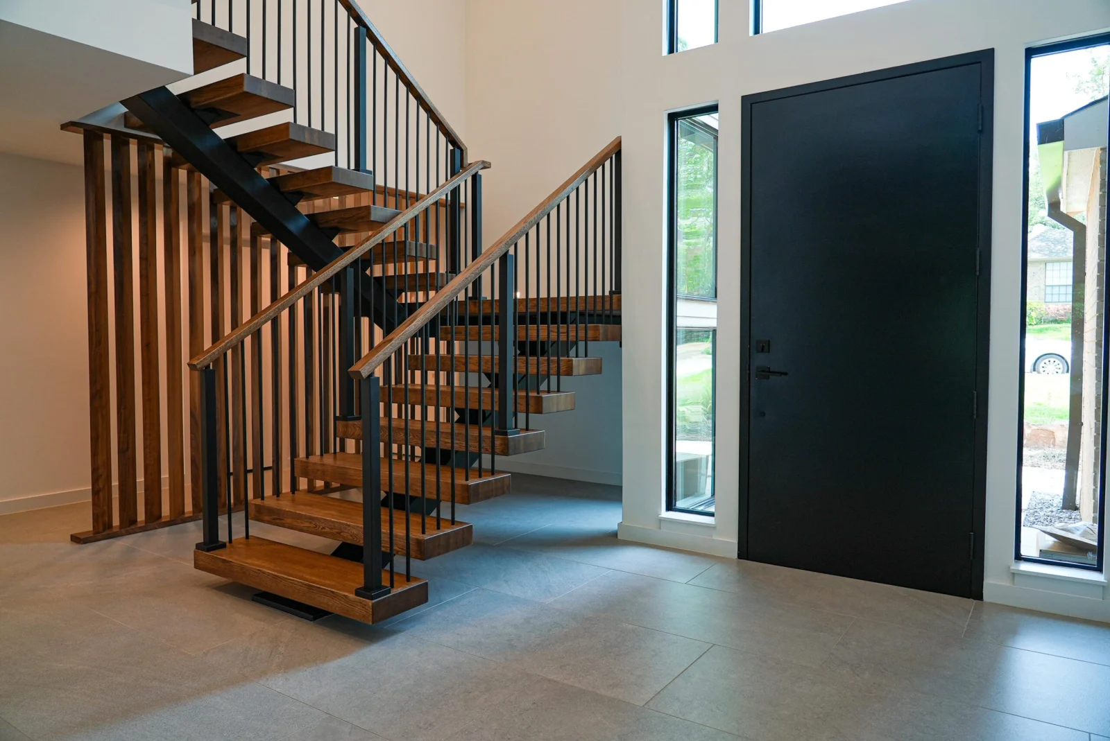 Medium wood treads on a black stringer with slim black balusters beside a black entry door