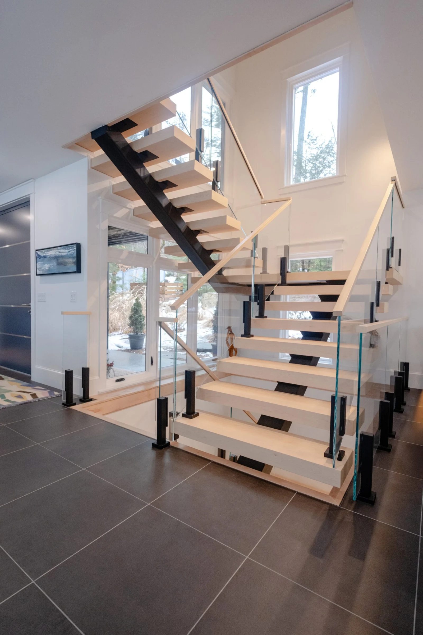 Switchback floating stair with pale treads, black glass clamps and a wood handrail over dark tile