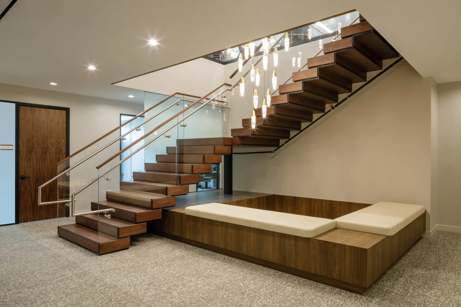 Stacked wood treads climb a lobby wall beside glass railing, a pendant cluster and built-in bench