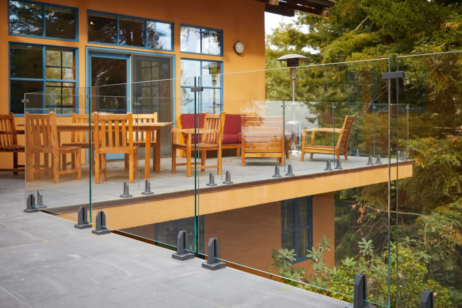Frameless glass panels on base clamps edge a tile deck with wood patio furniture and treetops