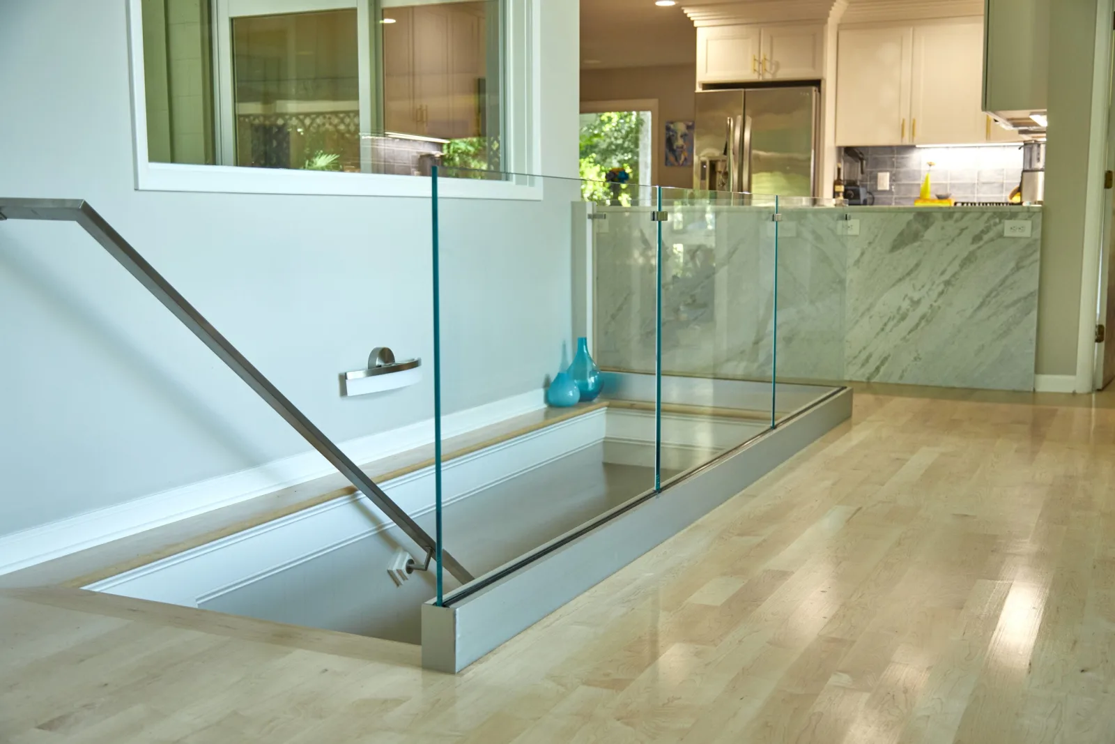 Glass railing on a metal base rail wraps a stair opening in a bright kitchen floor