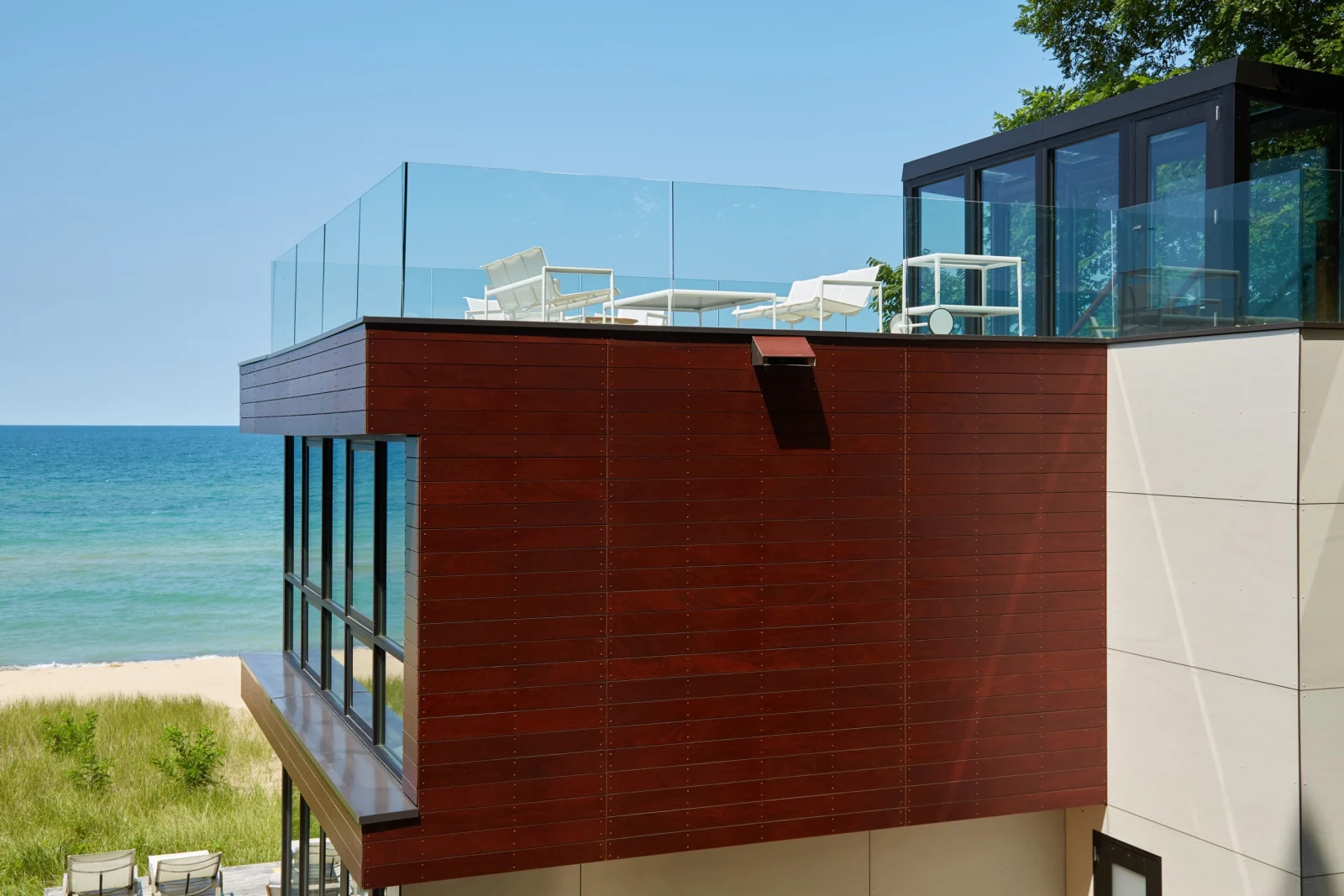 Rooftop deck ringed with glass railing on a red wood-clad house above a lakefront beach