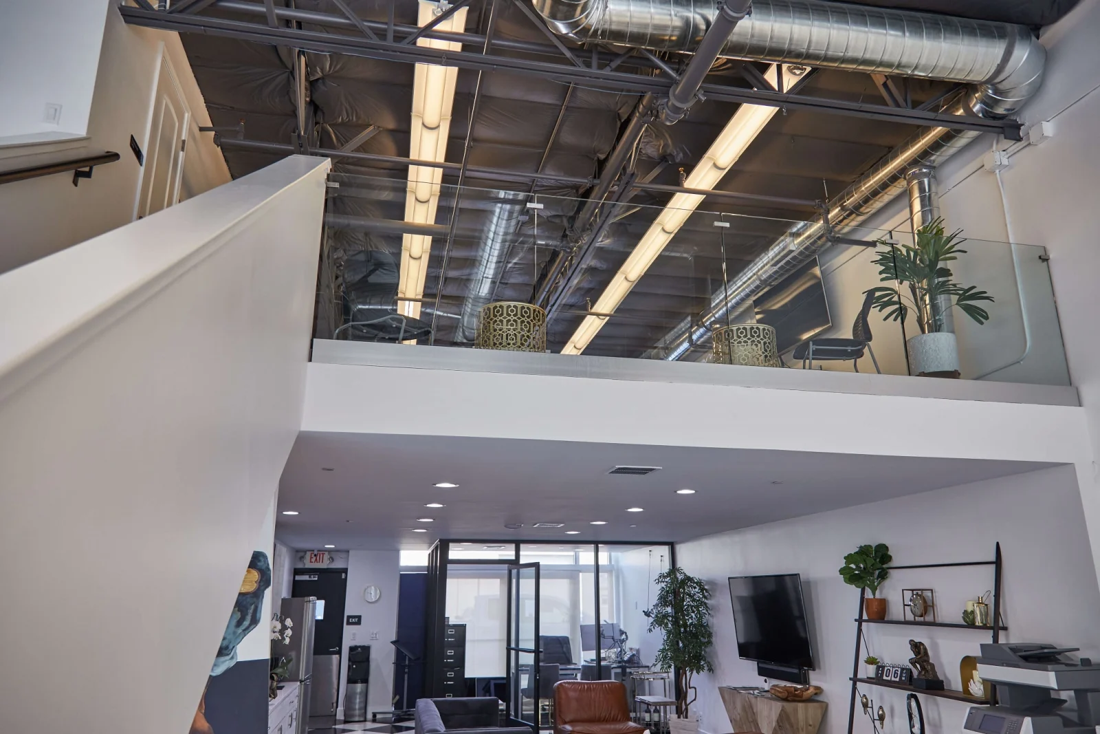 Glass railing along a loft mezzanine beneath exposed ducts and track lighting above an office lounge