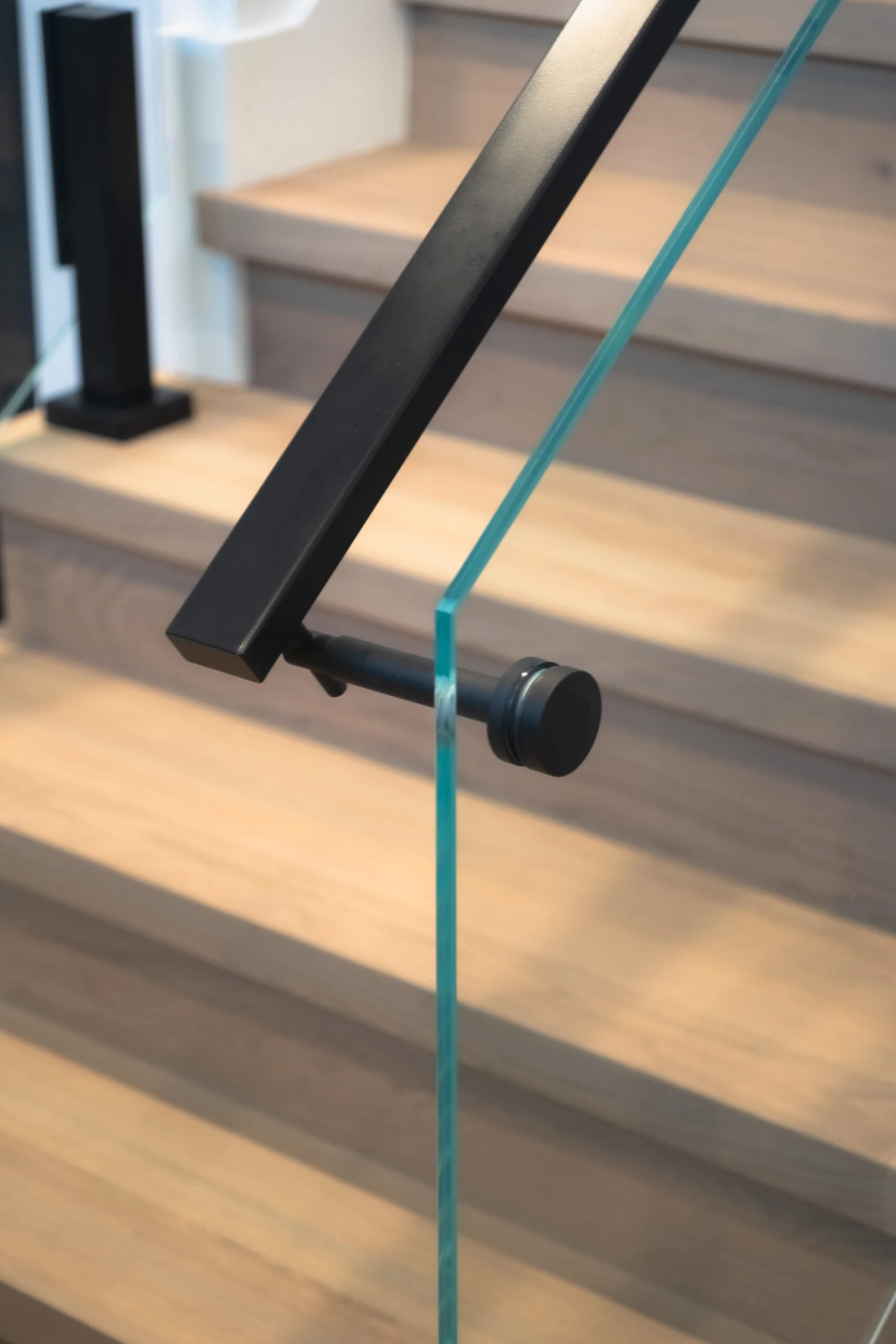 Black bracket clamps a glass panel to the stair handrail above wood treads