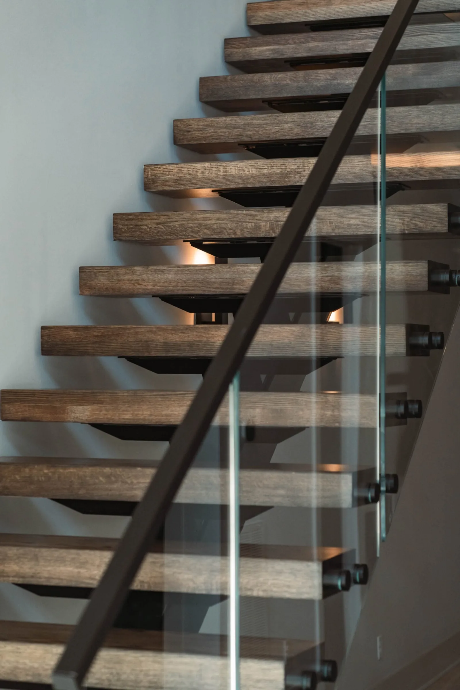 Gray-brown treads lit from beneath with glass panels on black standoff pins and a dark handrail