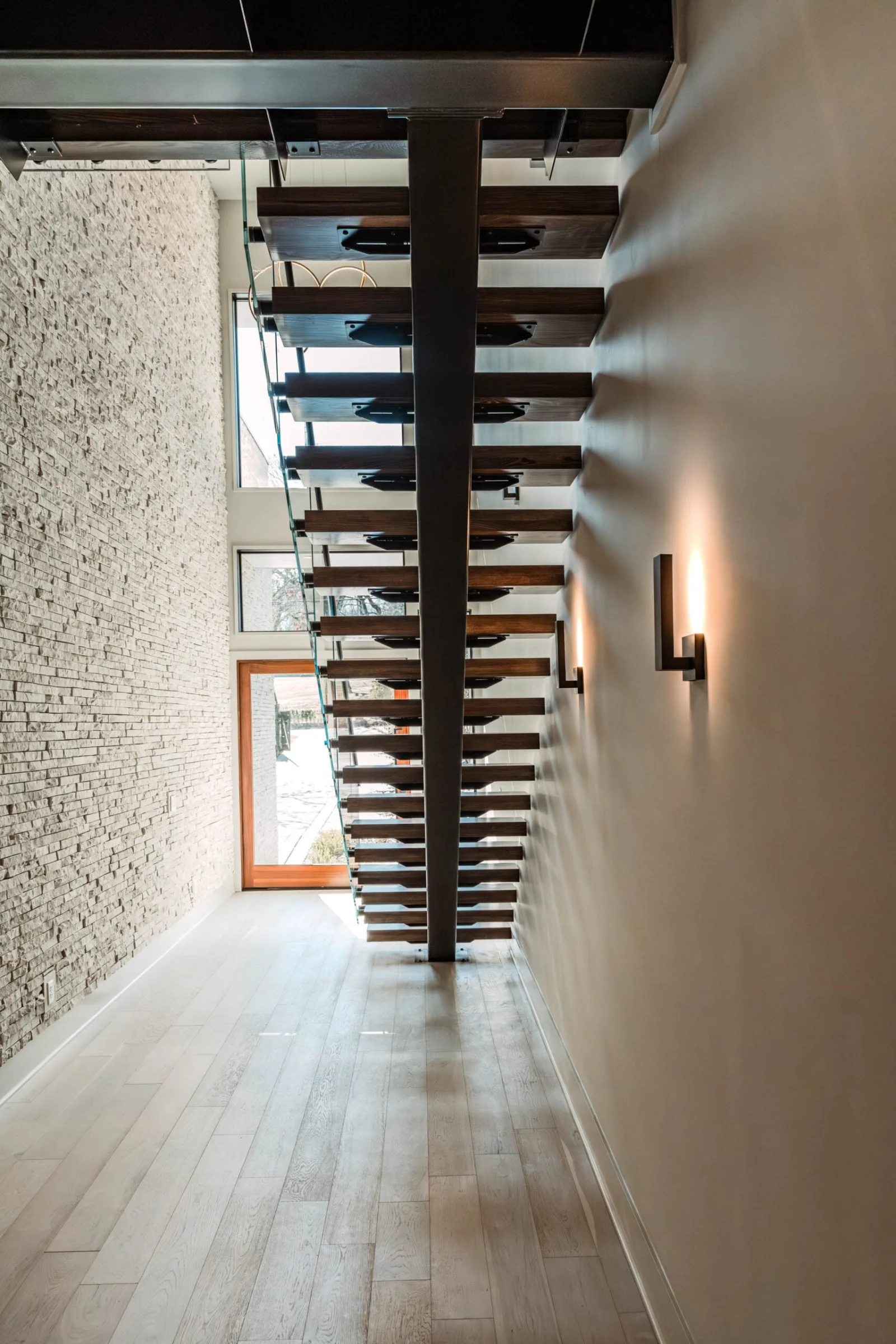 Corridor view of a floating stair rising between a brick wall and sconces