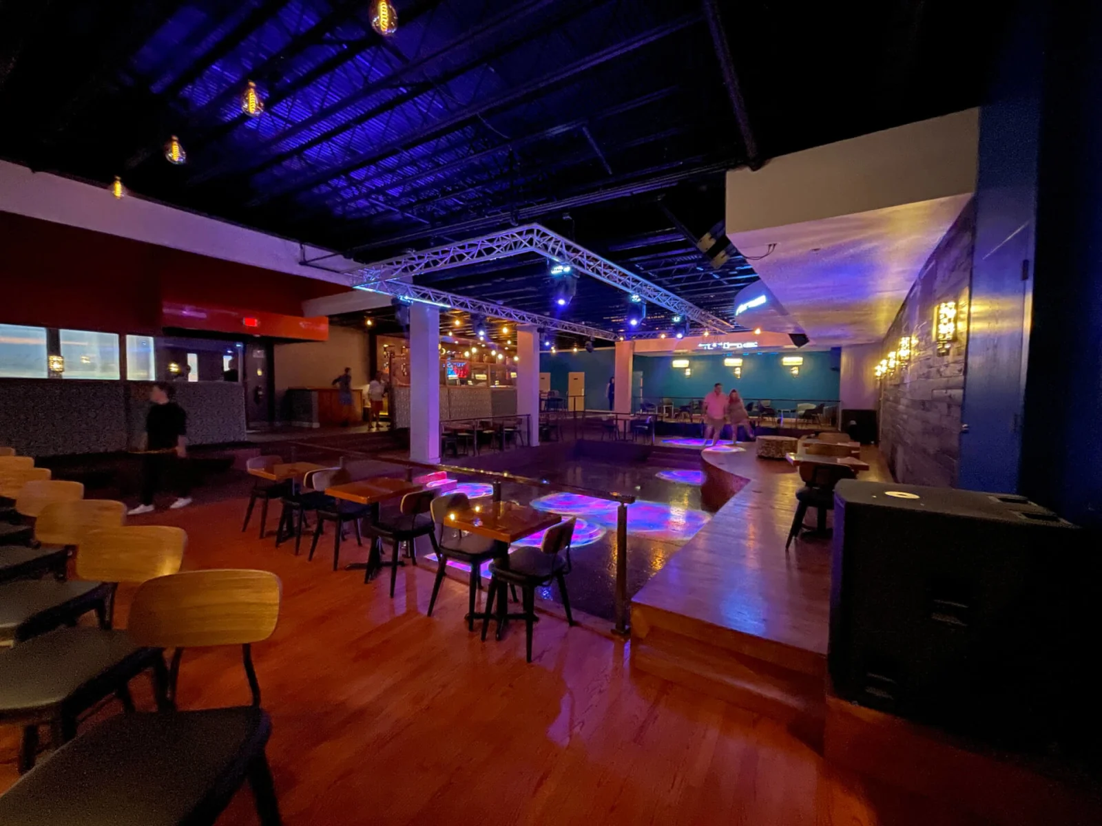 Nightclub interior under blue lighting with truss, tables and a black guardrail edging a platform