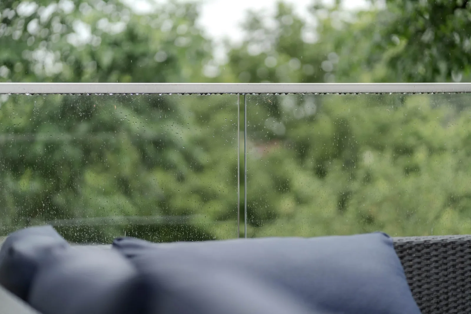 Rain-speckled glass panel and metal top rail seen close behind an outdoor cushion