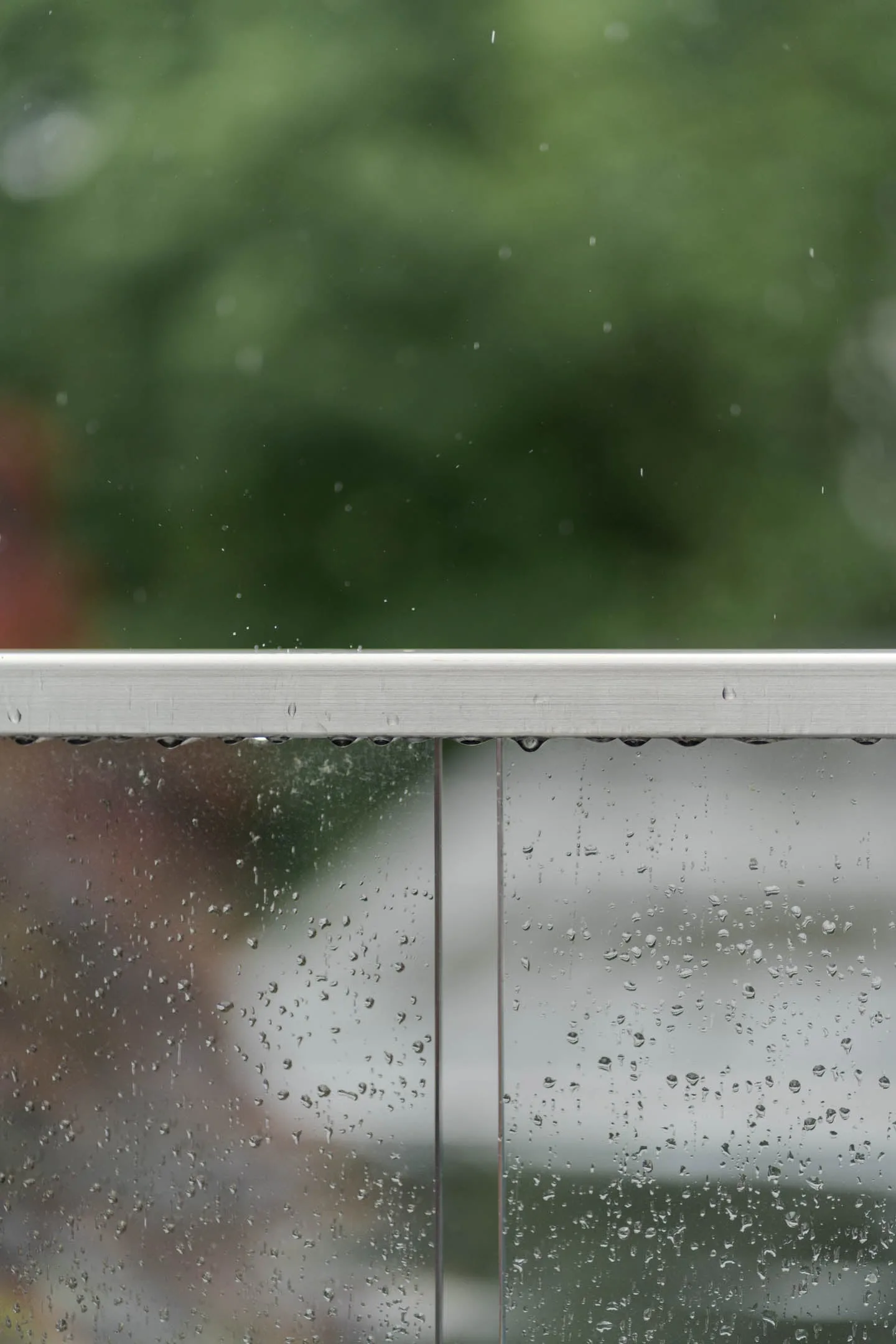 Rain-beaded glass panel under a metal top cap with blurred greenery beyond