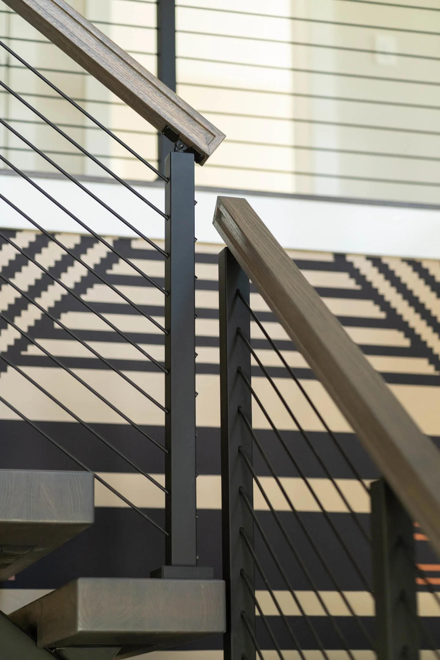 Black post and horizontal rod infill under a wood top rail, sunlight striping the wall