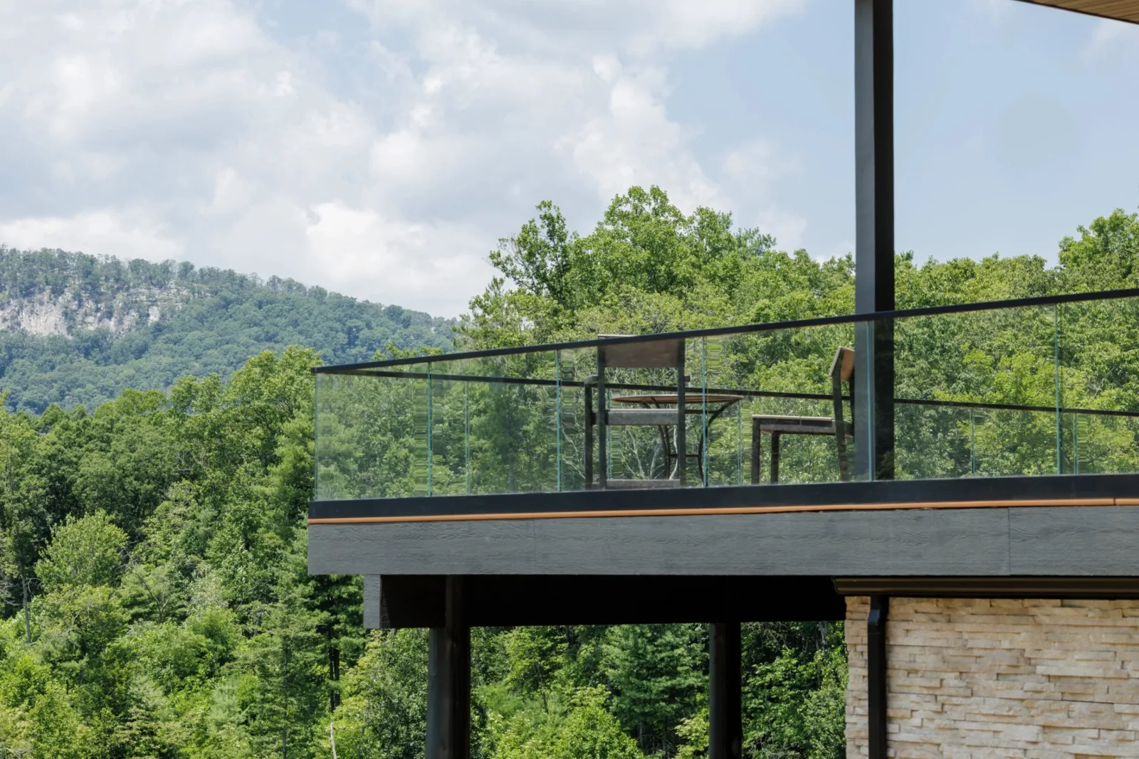 Cantilevered deck with glass railing juts over wooded hills on stone and steel supports