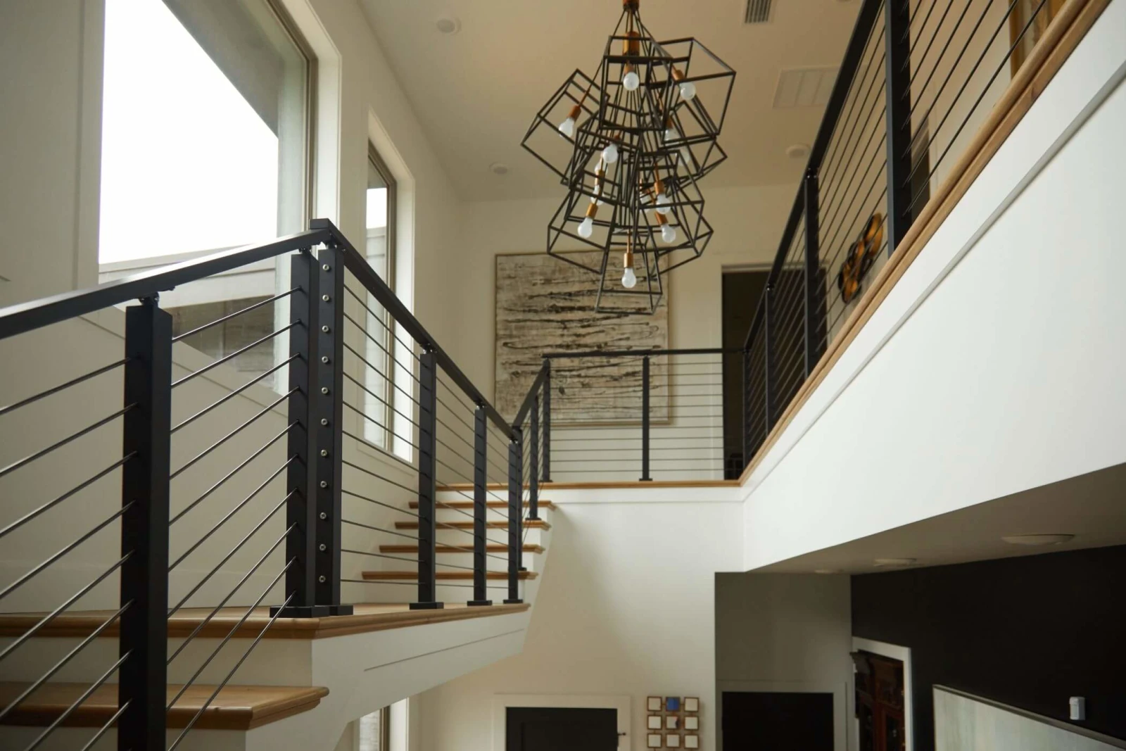 Two-story stairwell where black rod railing meets a wood cap under a caged light