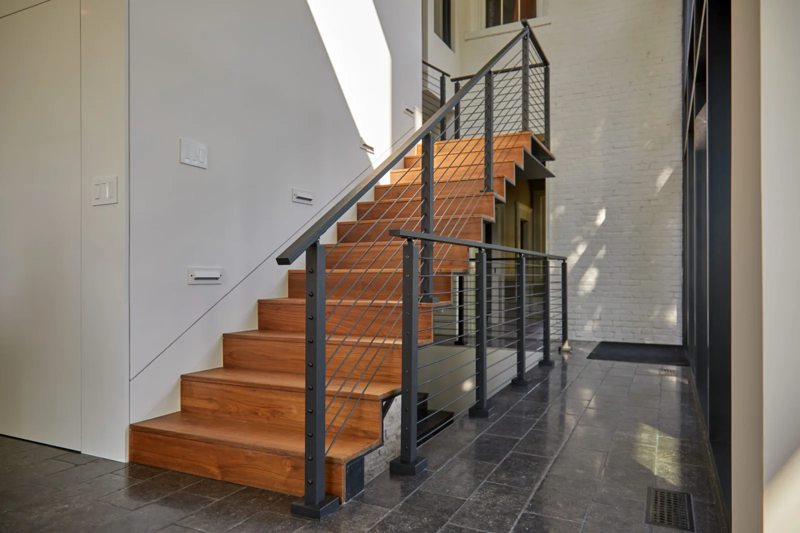 Straight wood stair with black rod railing rising from a tile entry beside painted brick