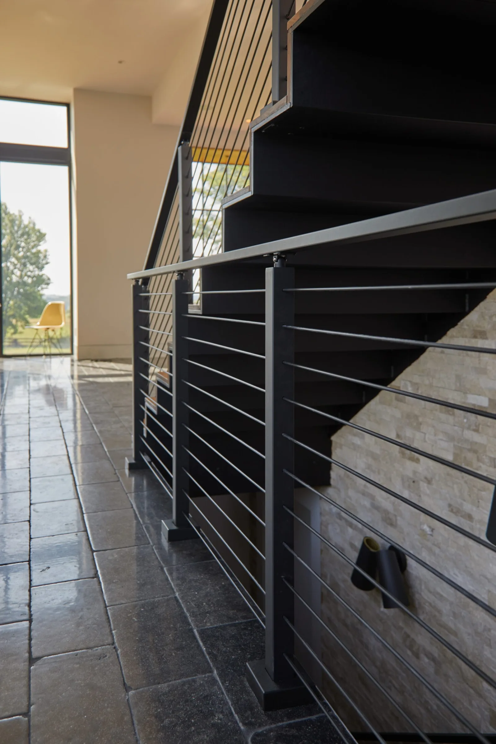 Black rod railing running beside a stone-tiled floor at the base of the stair