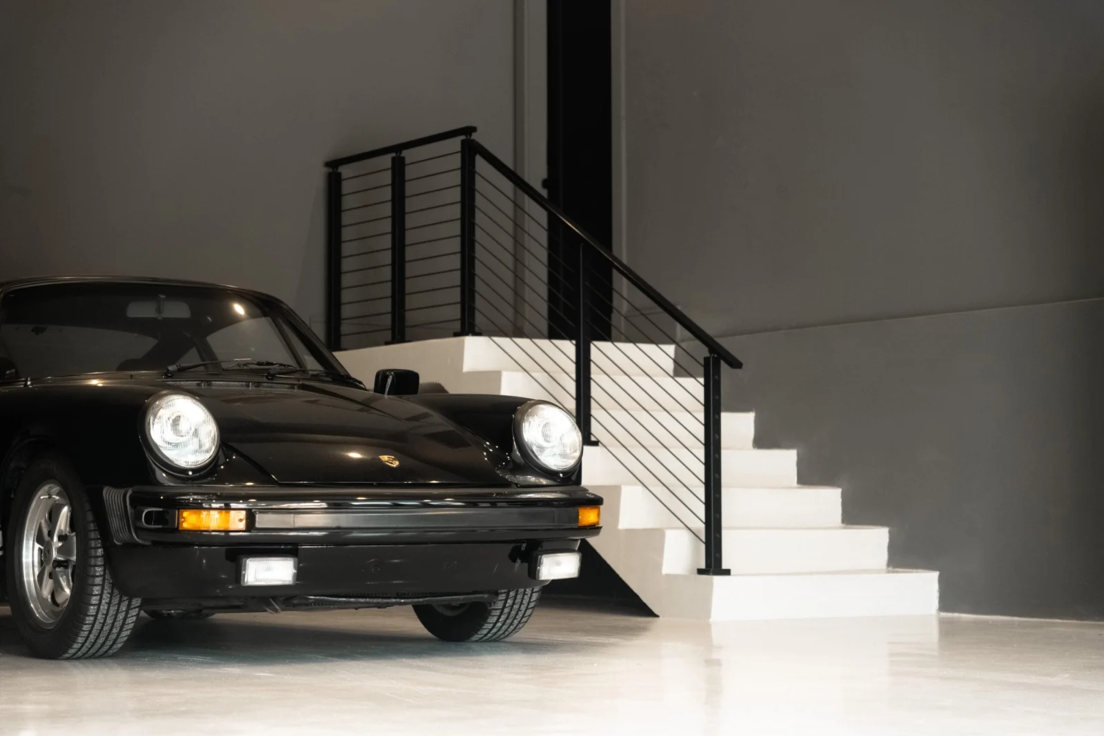 Black sports car parked beside white steps with a black rod railing in a showroom garage