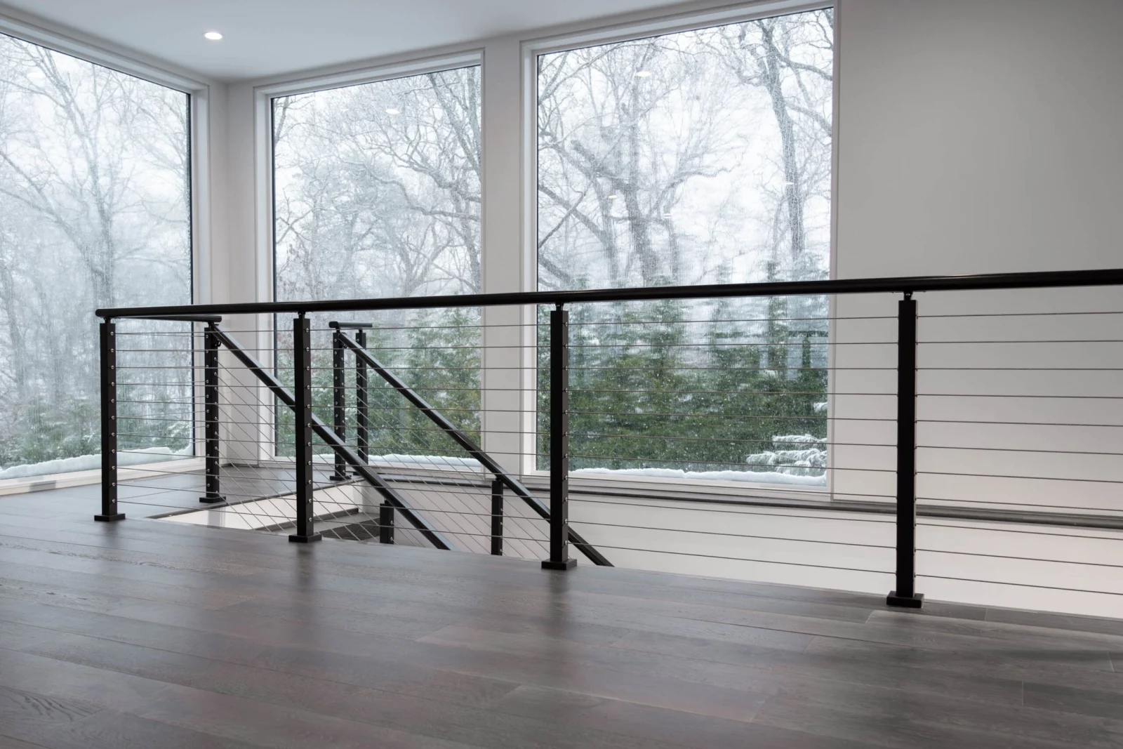 Cable railing guarding a stairwell opening in a bright room facing winter trees