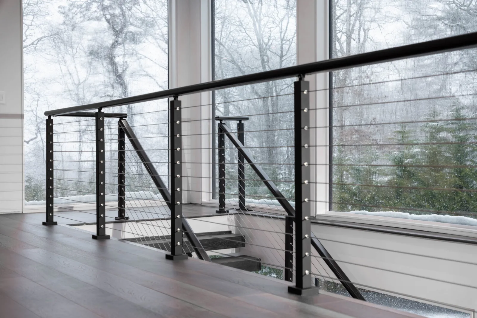 Black cable railing on a landing beside tall windows facing bare snowy woods