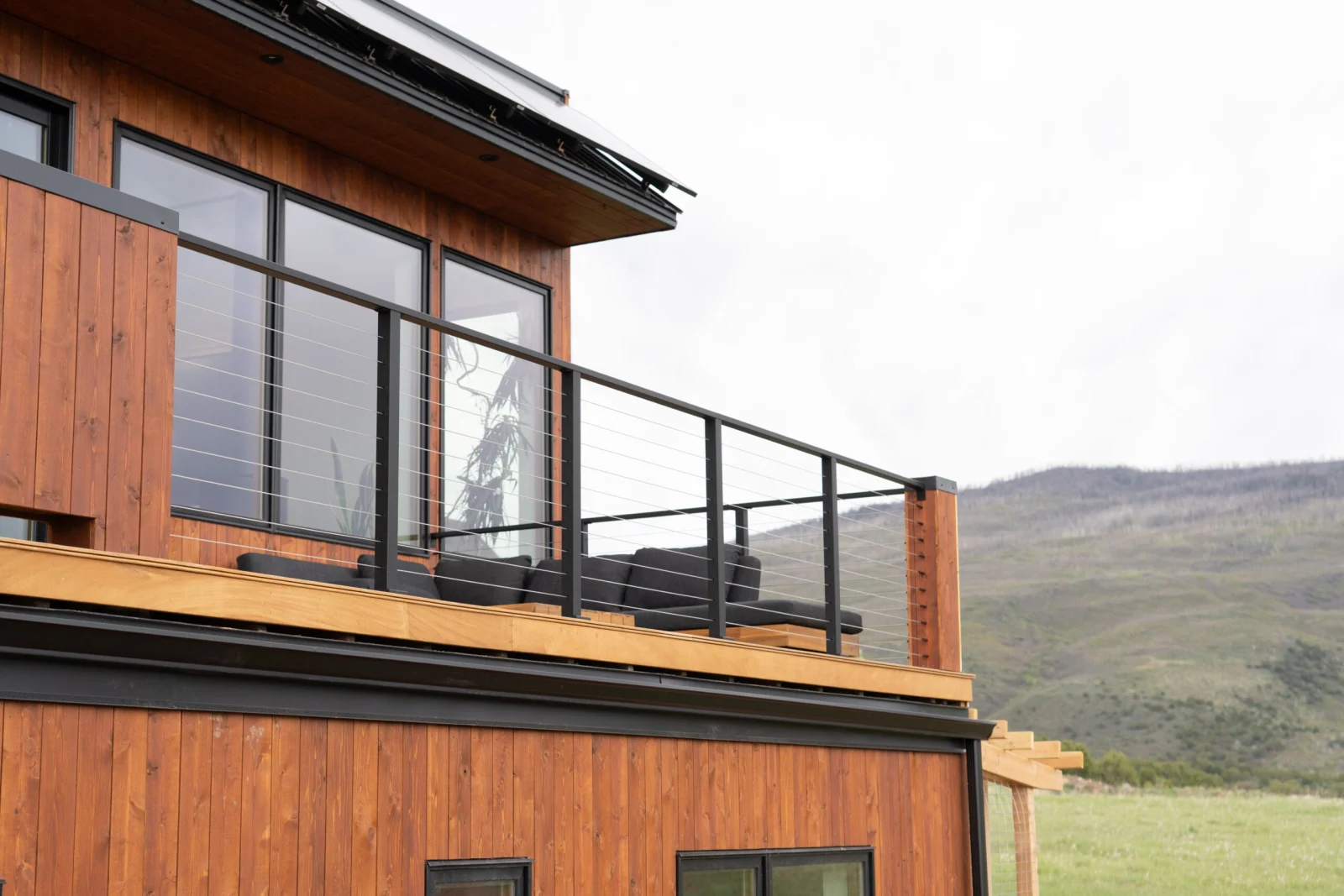 Wood-clad home with a black cable railing wrapping the upper balcony above open hills