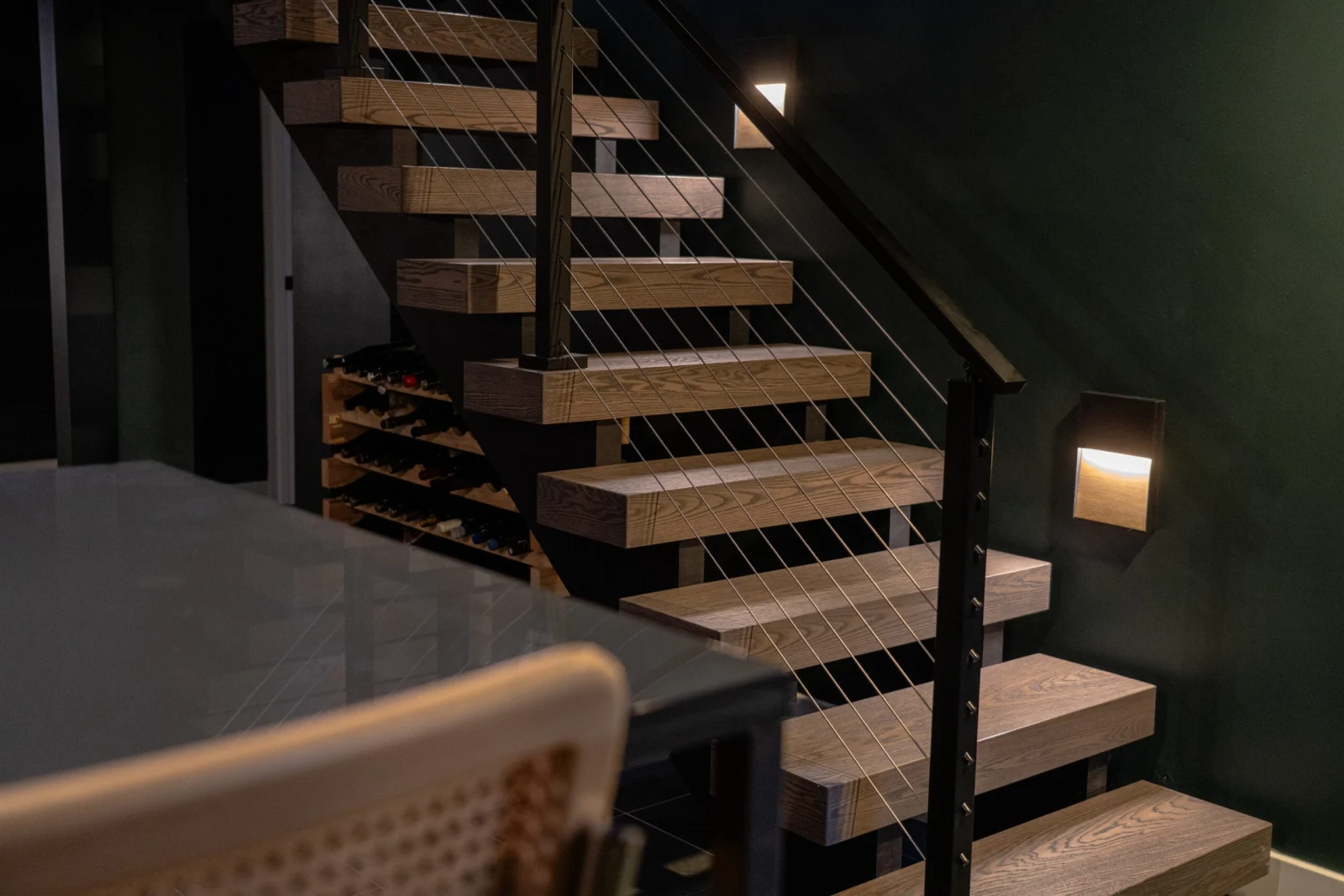 Stair with wood treads and cable railing above wine storage in a dark room