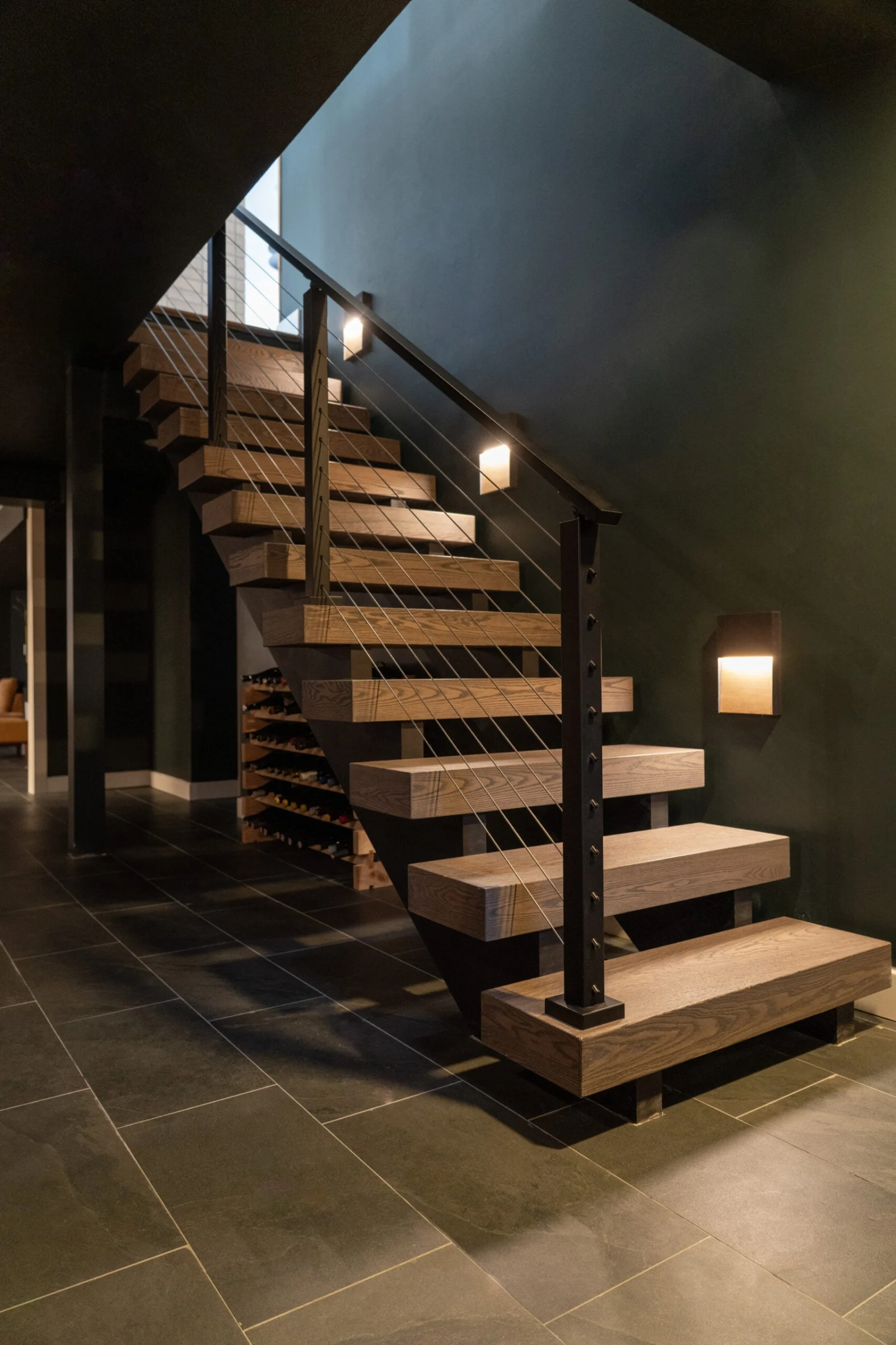 Dimly lit floating stair with thick treads and cable railing above a stone tile floor