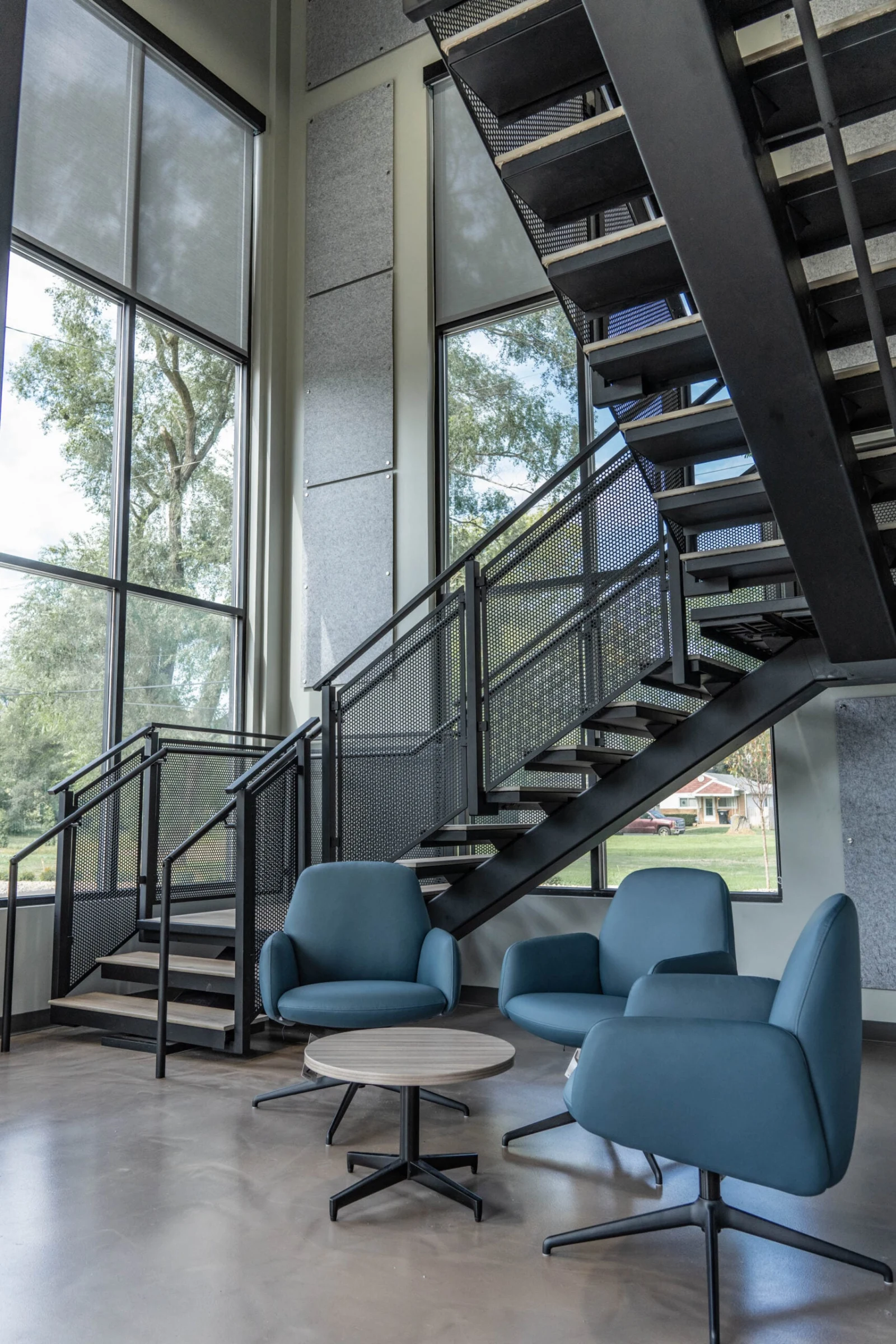 Tall window wall with the stair rising above blue lounge chairs and a round table