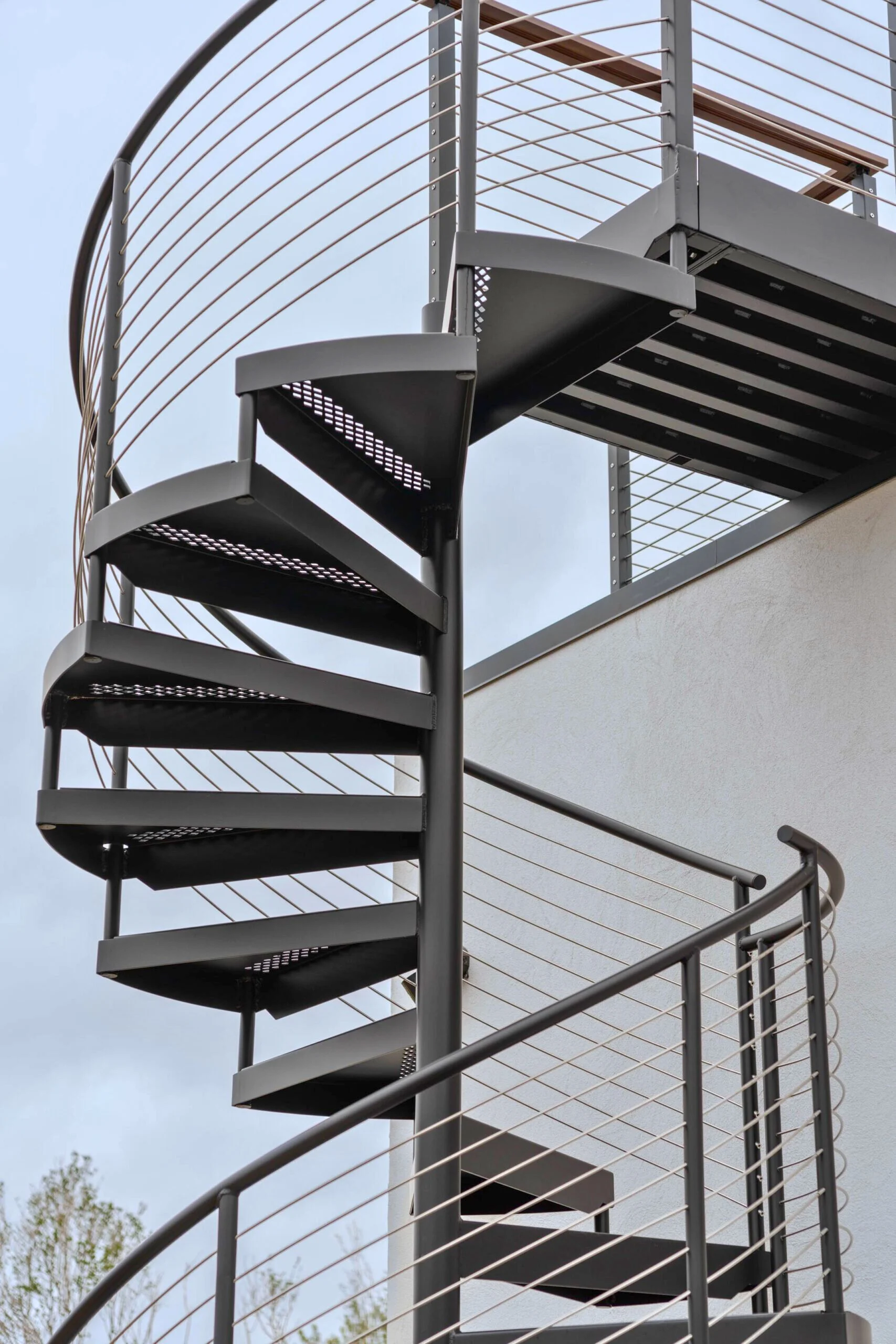 A black spiral stair with cable railing climbs against an exterior wall