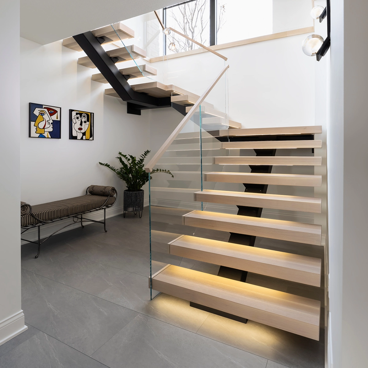Lit floating treads and a glass railing turn above a bench and framed prints