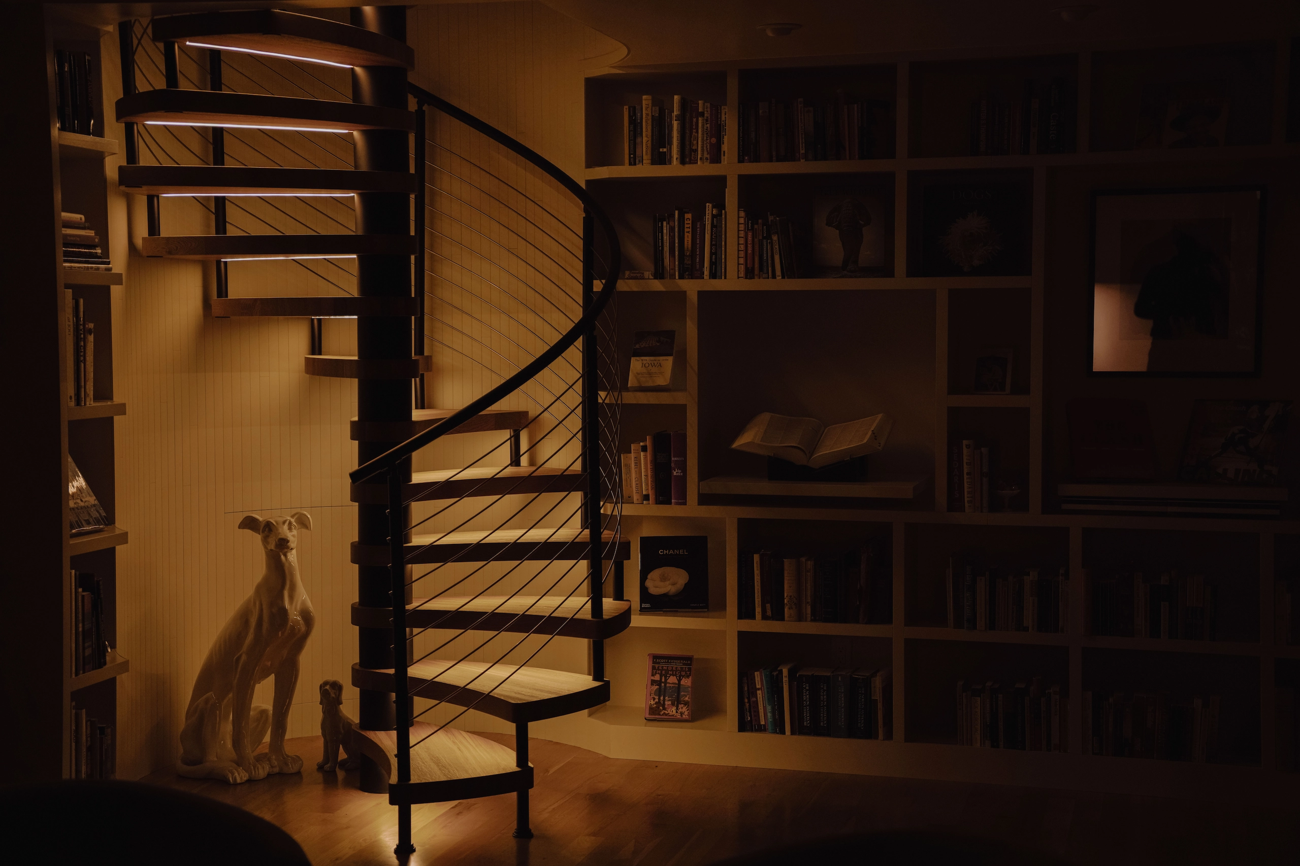 A spiral stair stands softly lit in a dim library corner beside a sculpture