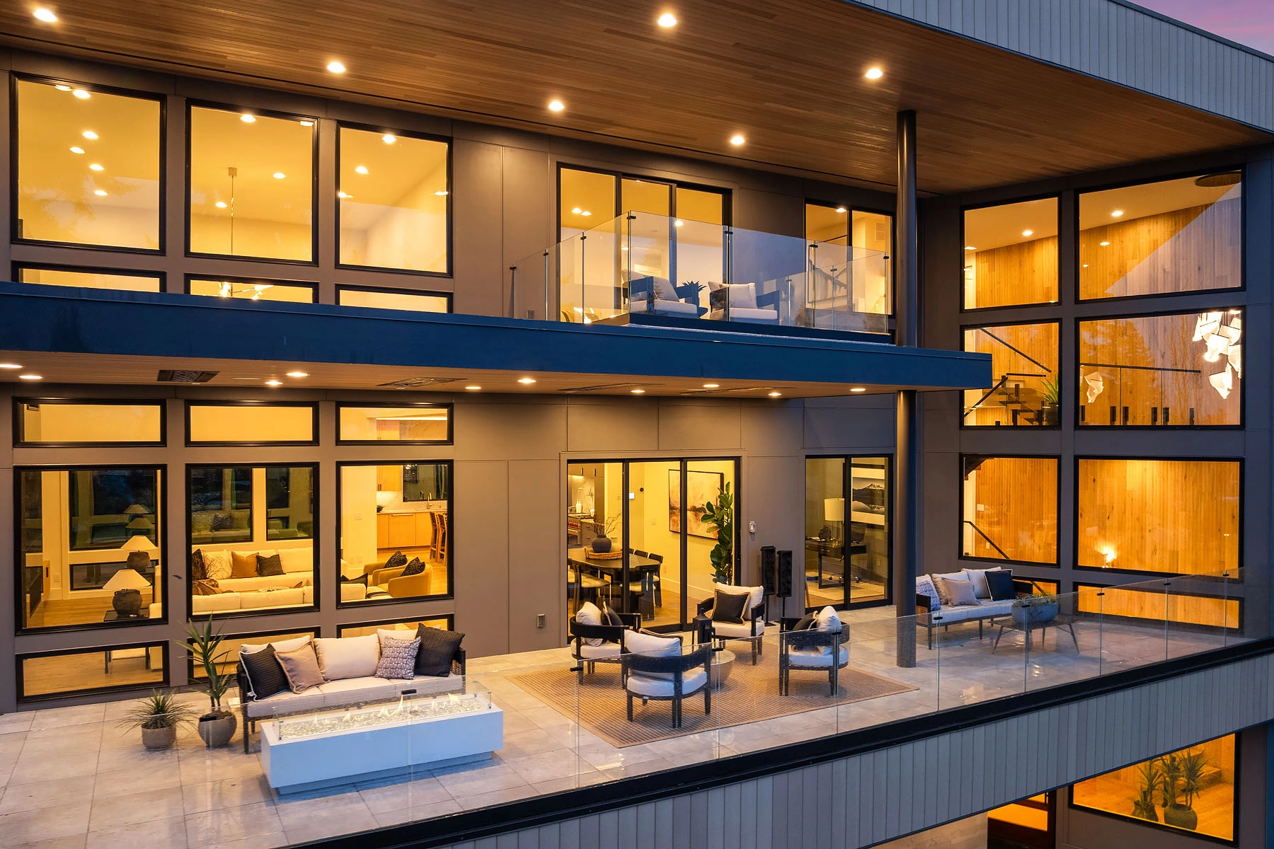 Glass railing wraps two levels of balconies on a home lit at dusk