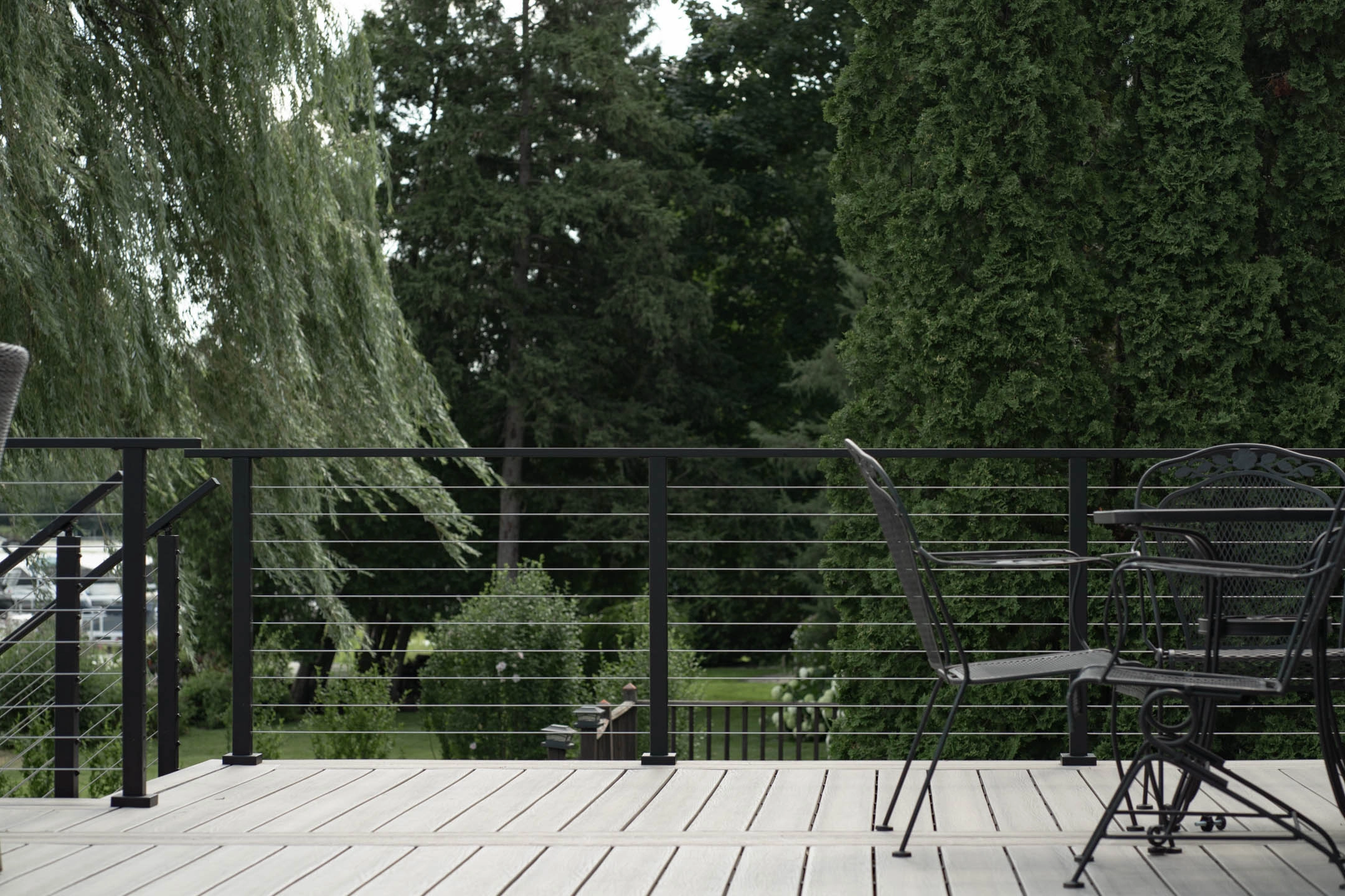 Black cable railing edges a deck with patio seating and a willow tree beyond