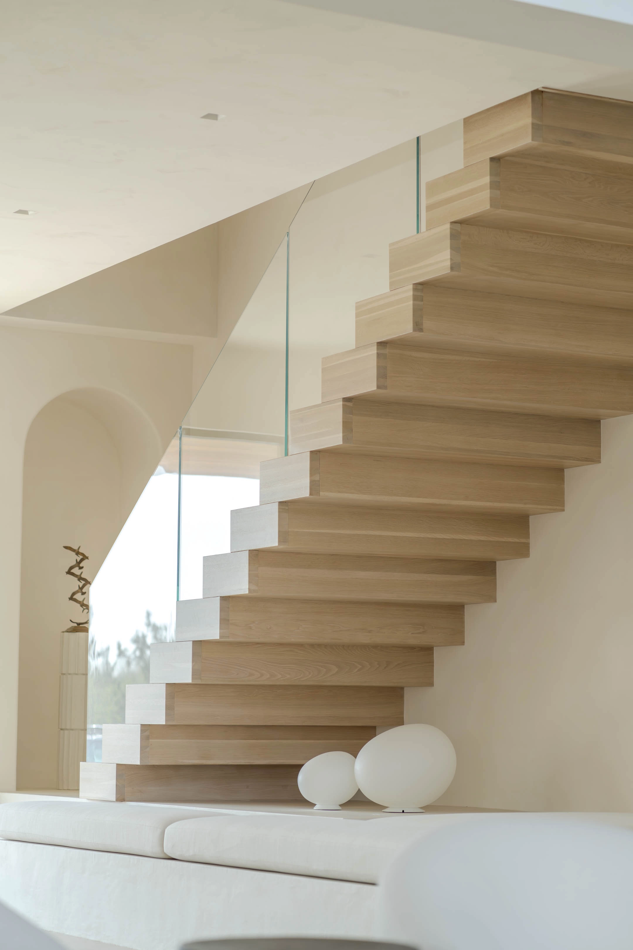 Stacked treads climb a bright stairwell behind a frameless glass railing