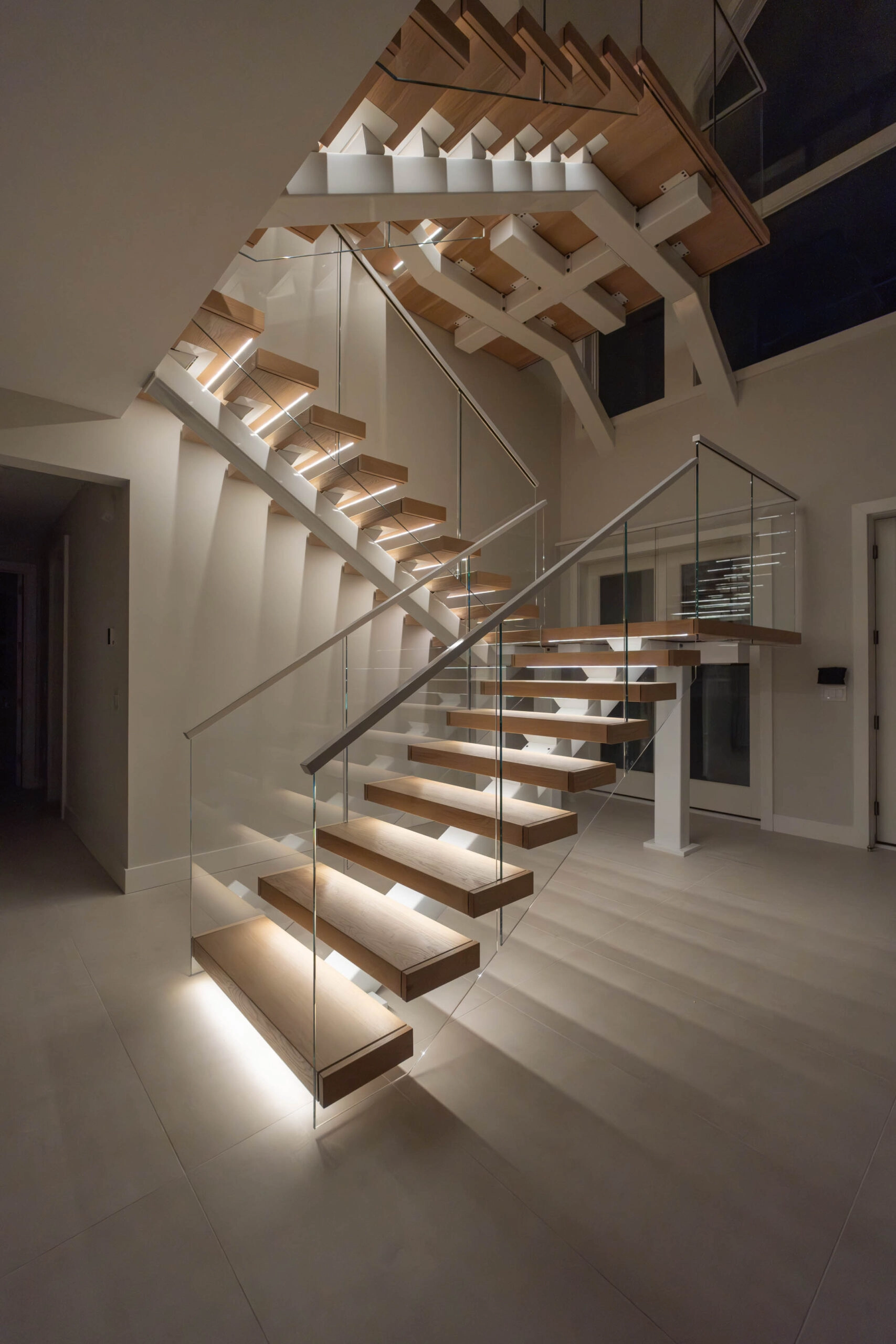 Lit treads climb a multi-story stairwell behind frameless glass at night