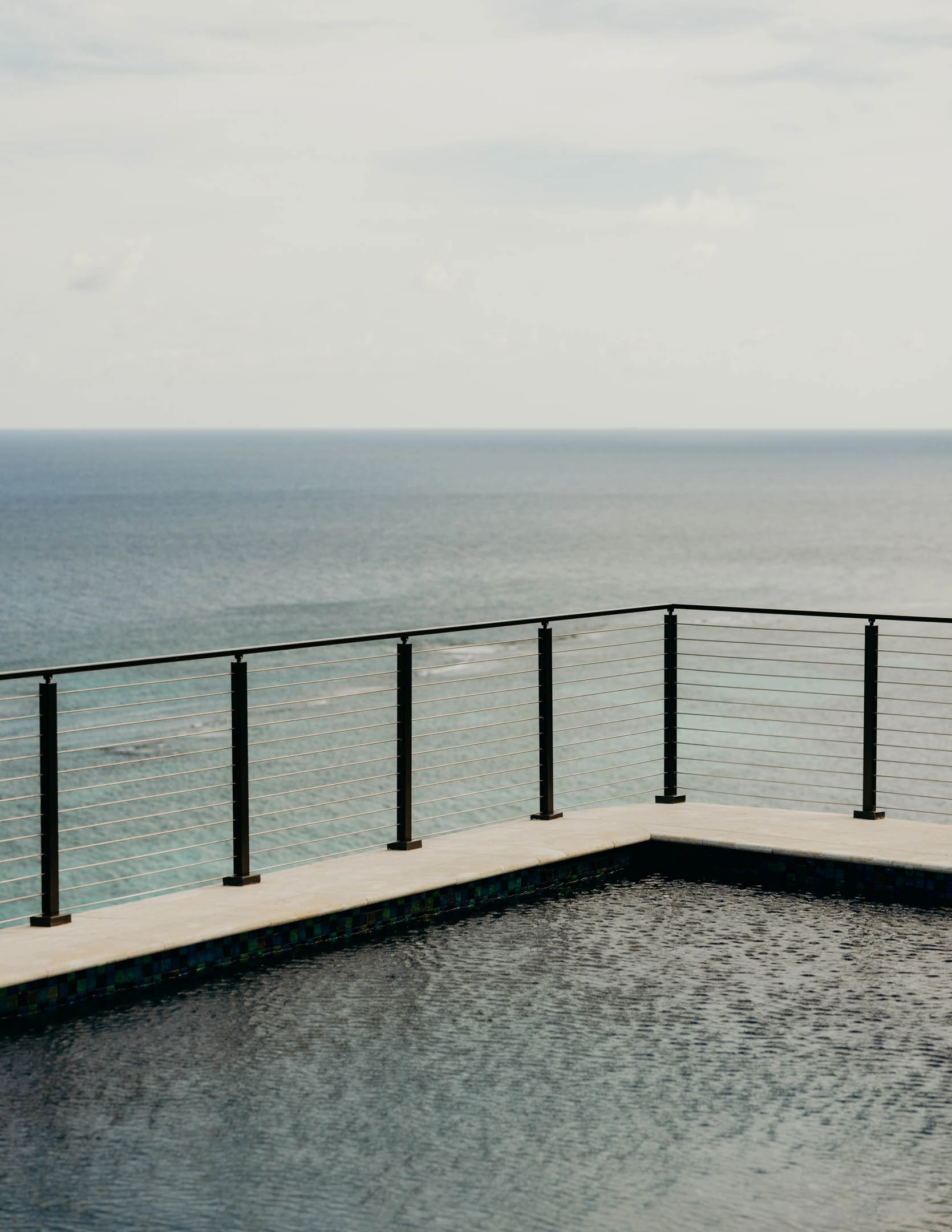 Rod railing follows a cliff-edge pool deck above a still grey sea