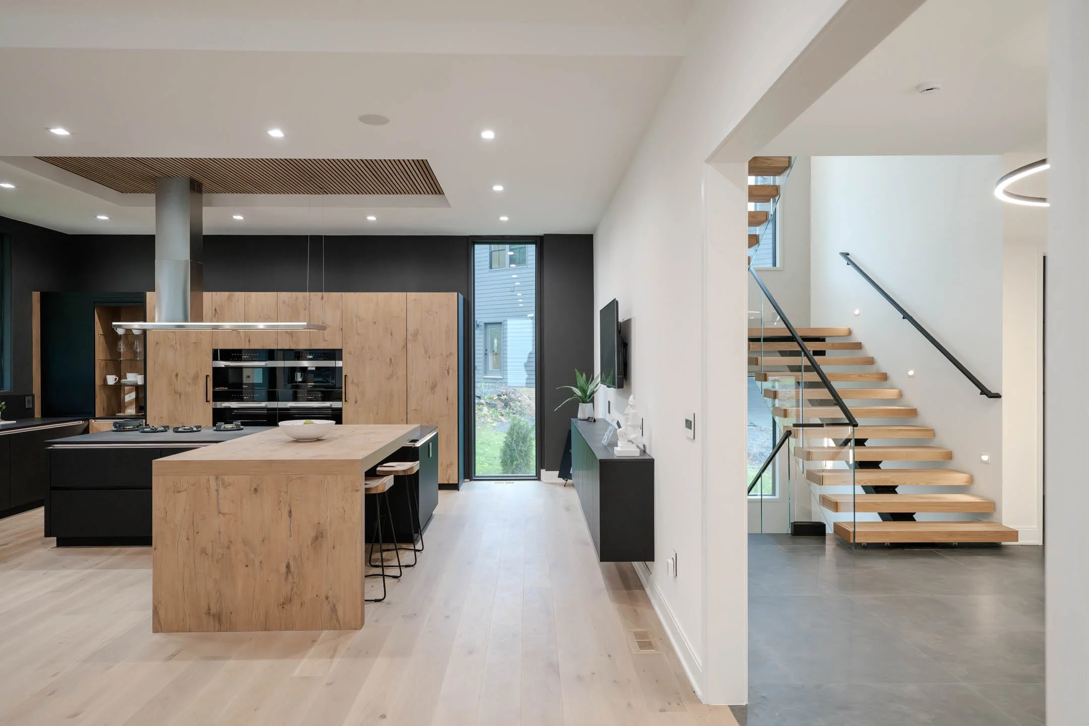 A floating stair with glass railing stands past a dark open kitchen