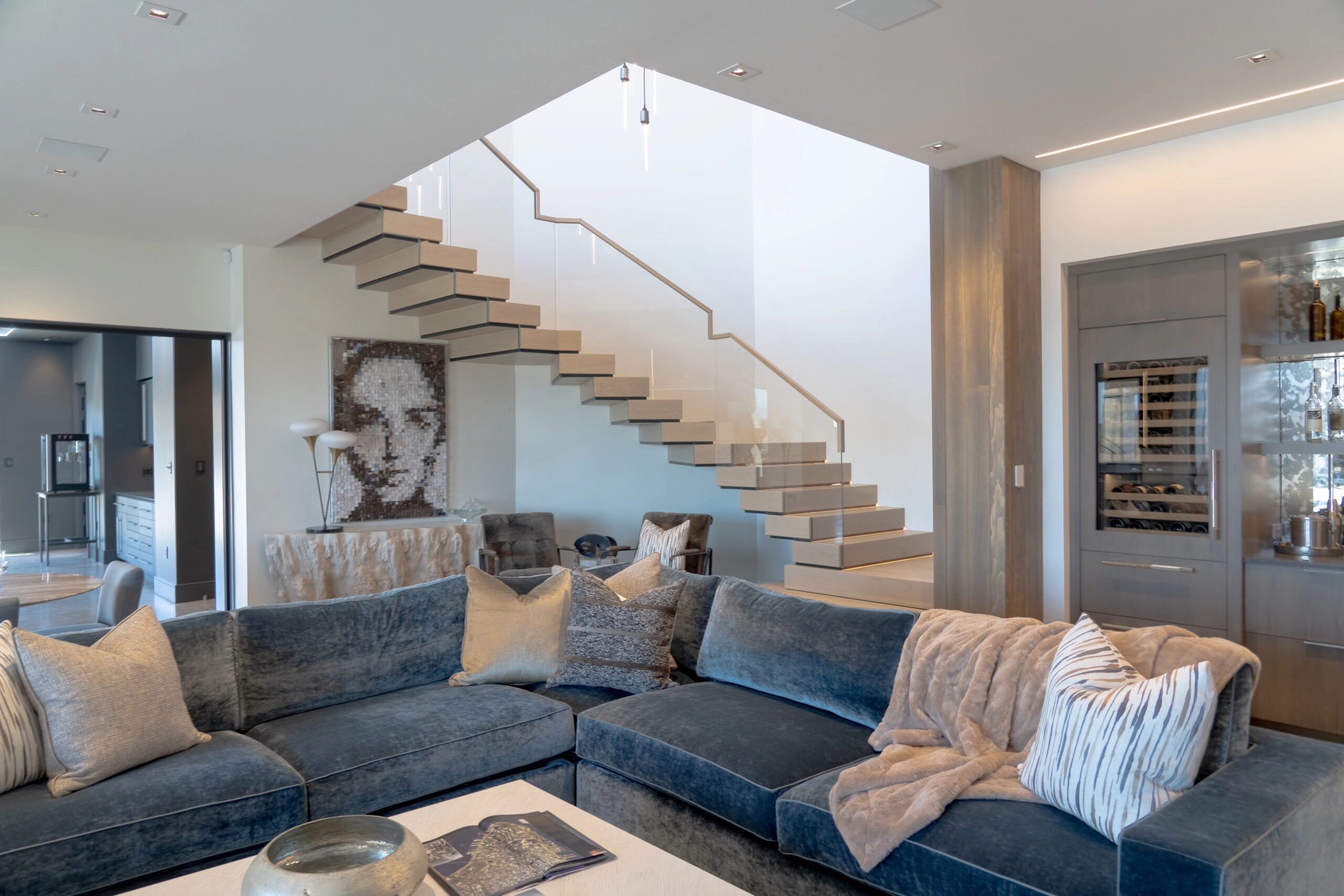 A stacked stair climbs behind a deep sectional in a soft, bright living room
