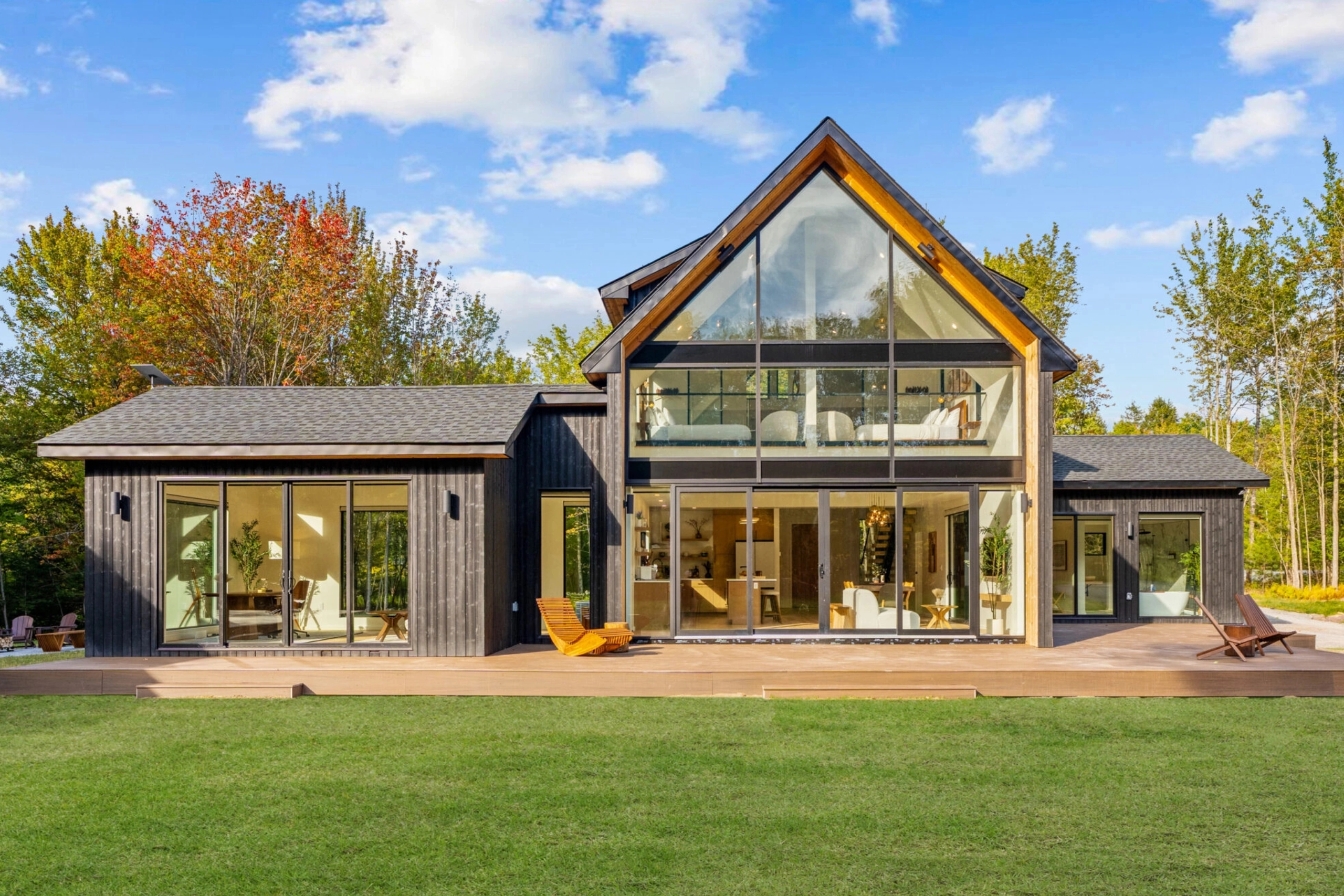 A gabled house wall of glass faces a wide lawn under a bright blue sky