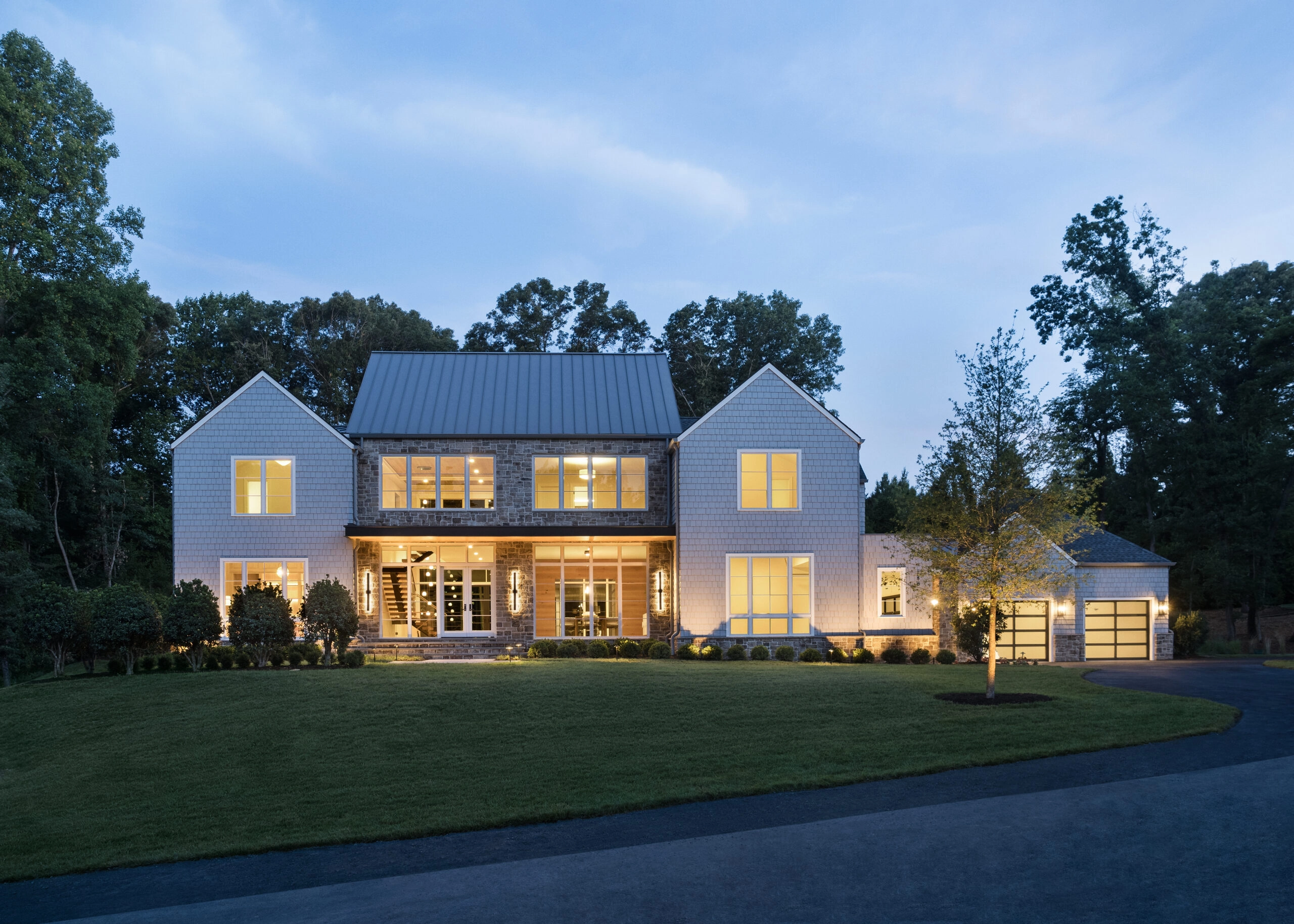 A large home glows at dusk behind a broad lawn and mature trees