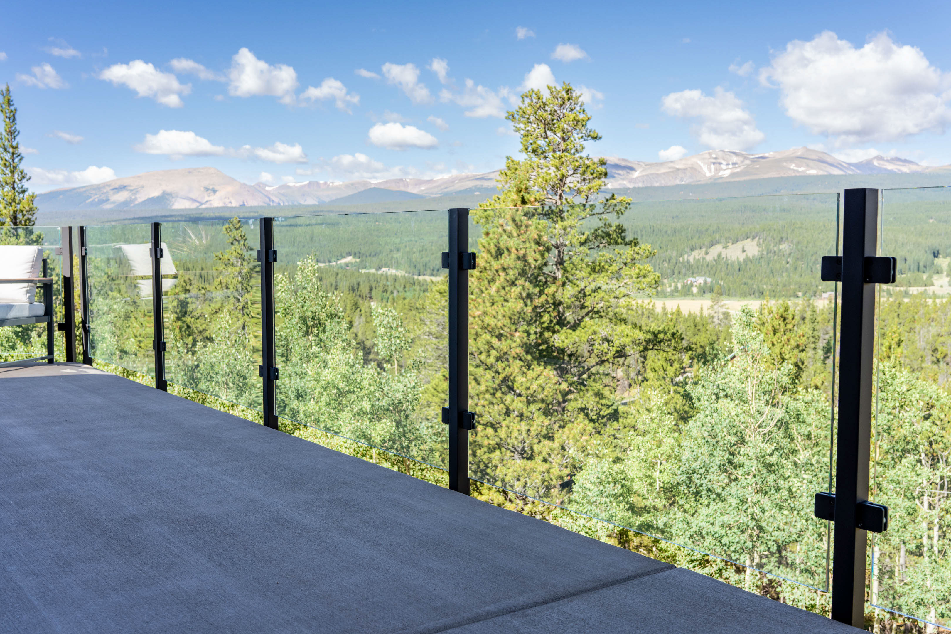 A glass and black post railing lines a deck above pines and distant peaks